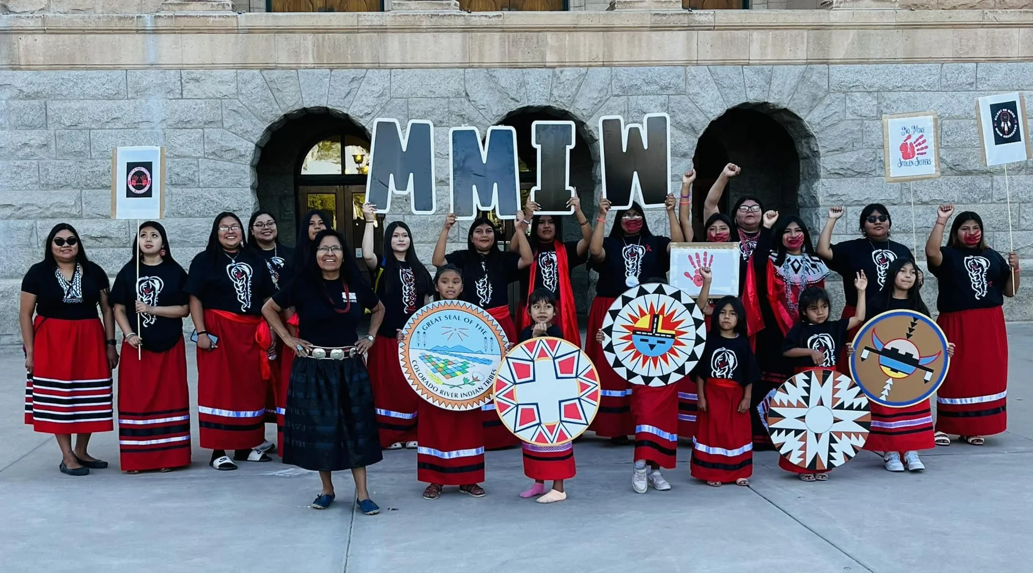 Group of women and children in traditional Native American clothing holding signs and symbols, standing in front of a stone building with large letters spelling 'MLTW' and celebrating Indigenous culture.