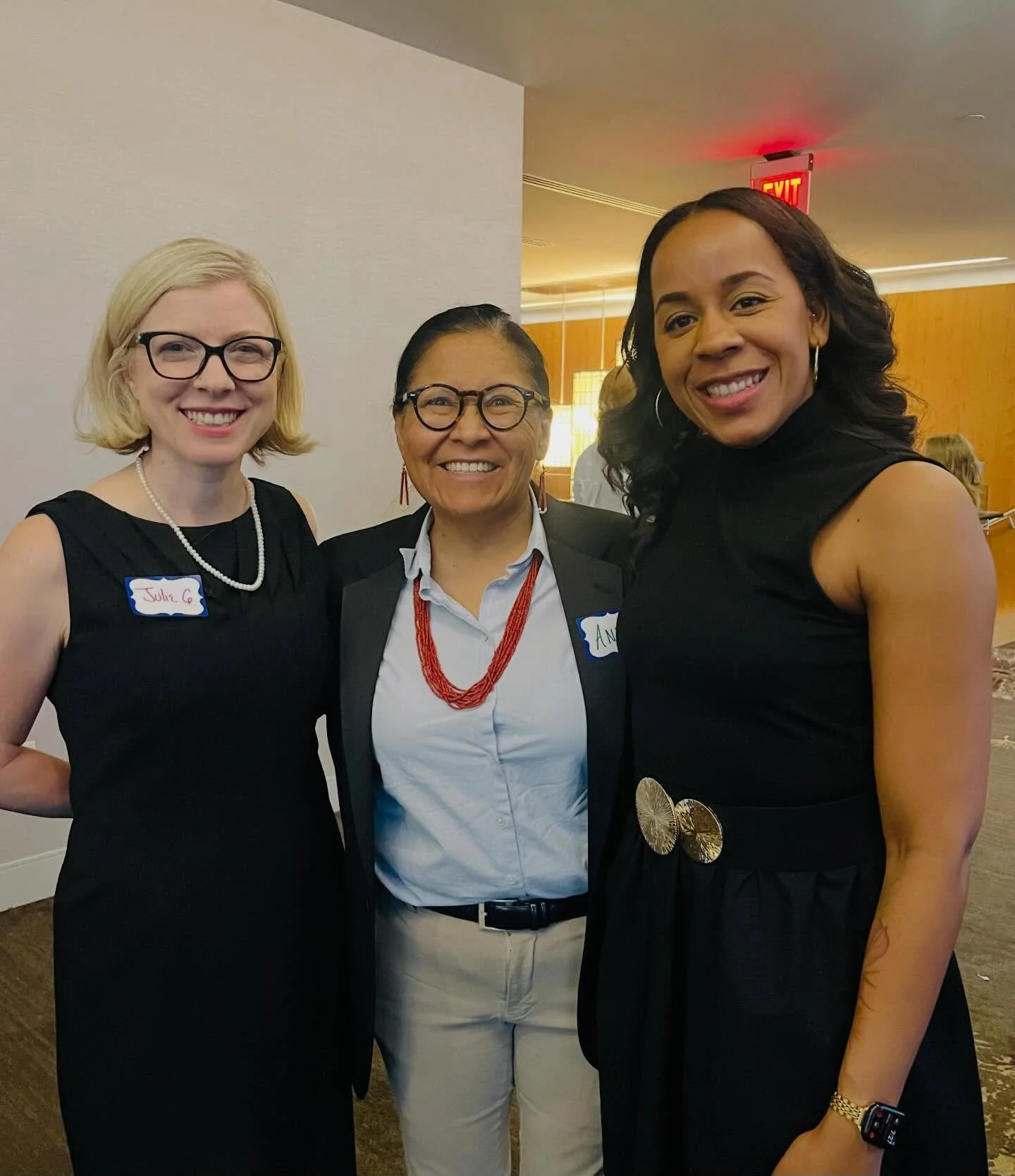 Three women standing together and smiling at an indoor event, wearing business attire, with two women on the left and right wearing black dresses and the woman in the middle wearing a white blouse and blazer.