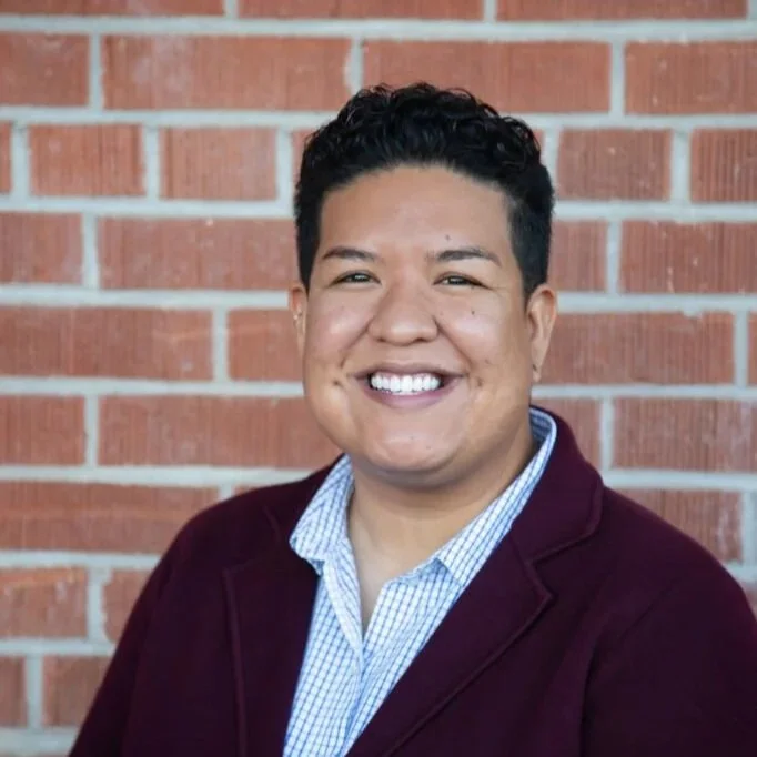Portrait of a smiling man with dark, curly hair, wearing a blue checkered shirt and a maroon blazer, standing in front of a brick wall.