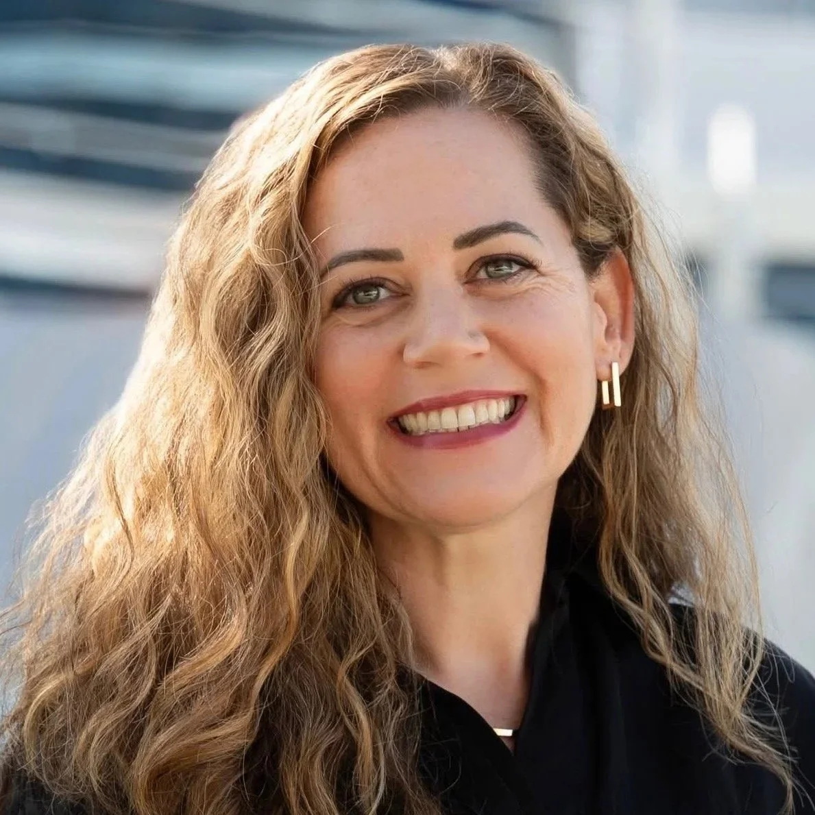 A woman with wavy, light brown hair and blue eyes, smiling at the camera, wearing a black top and gold earrings, outdoors with a blurred cityscape background.