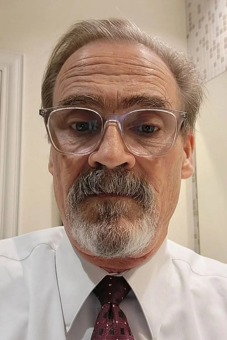 A middle-aged man with glasses and a beard taking a selfie in an indoor setting, wearing a white shirt and a dark red tie.