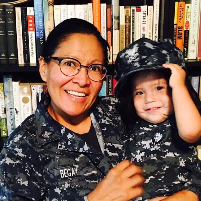 Woman in military uniform with glasses holding a smiling young child in camouflage outfit and cap, standing in front of a bookshelf filled with books.
