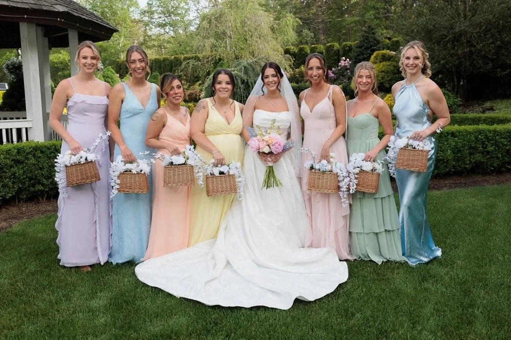 Bride surrounded by seven bridesmaids outdoors, all dressed in pastel-colored gowns, holding baskets with white flowers, with green trees and bushes in the background.