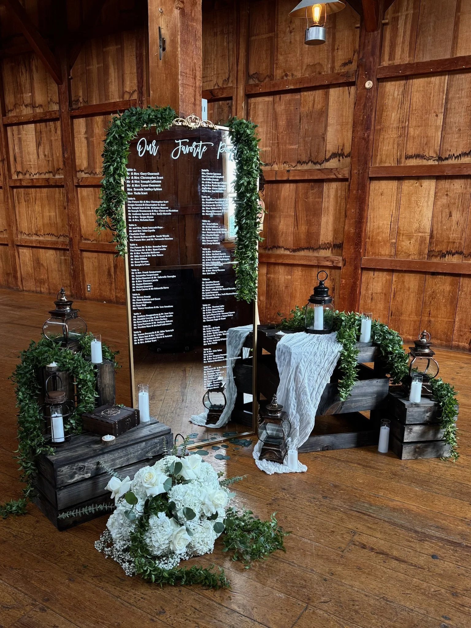 Rustic indoor wedding or event decor with a black tabletop seating chart, surrounded by greenery, candles, and white flowers, set against wooden walls.