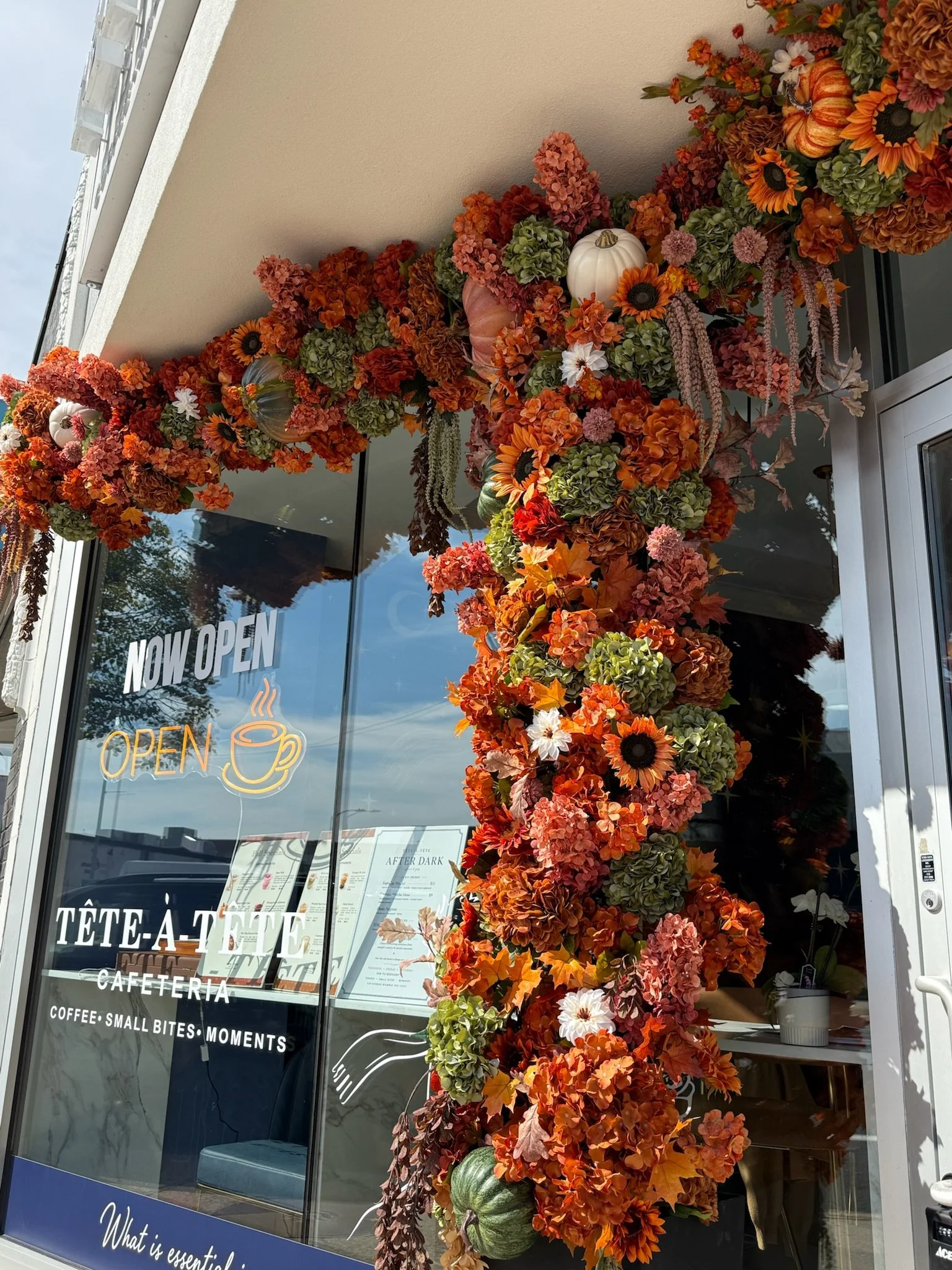 Autumn-themed decoration with orange, green, and white flowers, pumpkins, and foliage outside a cafe with glass window displaying 'Now Open' sign.