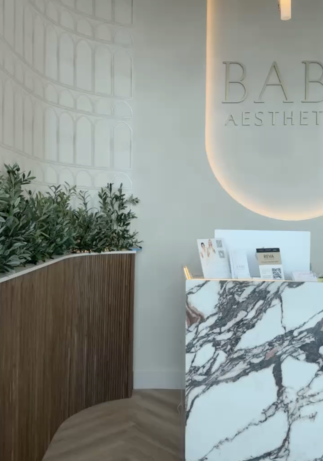 Modern reception area with green plants in a wooden planter, a marble reception desk, and a wall with the sign 'BABY AESTHETICS' illuminated by backlighting.
