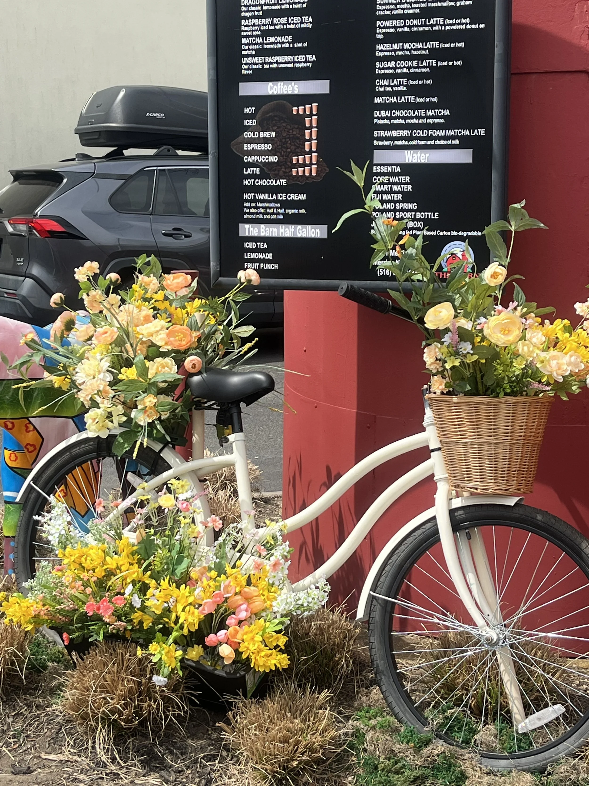 A white bicycle with a wicker basket filled with colorful flowers, positioned next to a large outdoor menu board and situated in front of a red wall, with a black car parked behind.