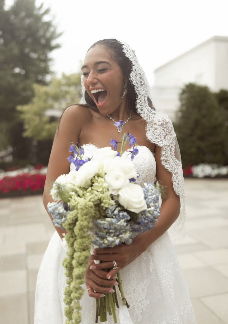 A smiling bride in a white lace wedding dress holding a bouquet of white and purple flowers, outdoors with trees and a building in the background.