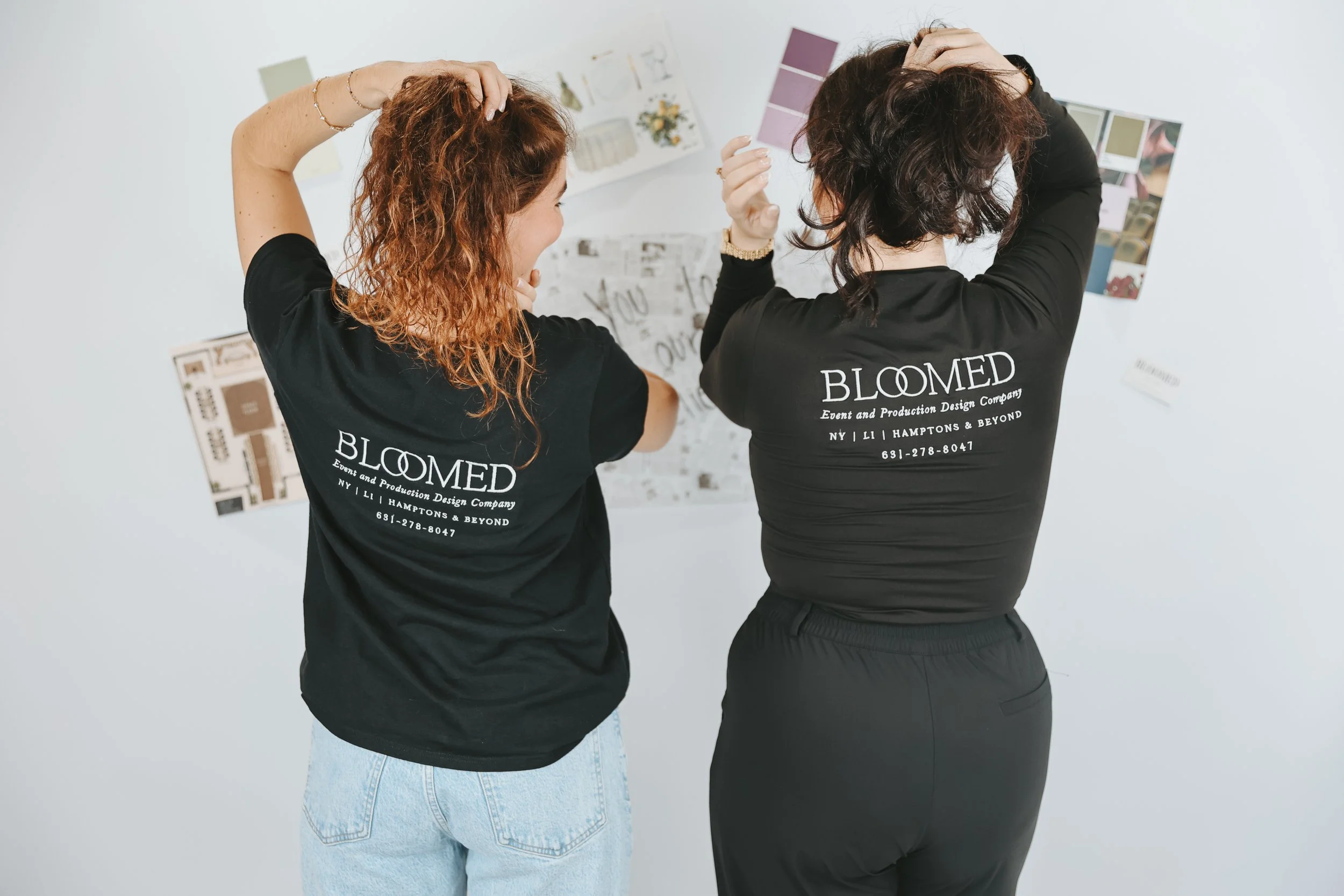 Two women standing with their backs to the camera, wearing matching black T-shirts with white text that reads 'BLOOMED Event and Production Design Company NY LI HAMPTONS & BEYOND 631-278-8047'. The women are touching their hair and looking at a wall 