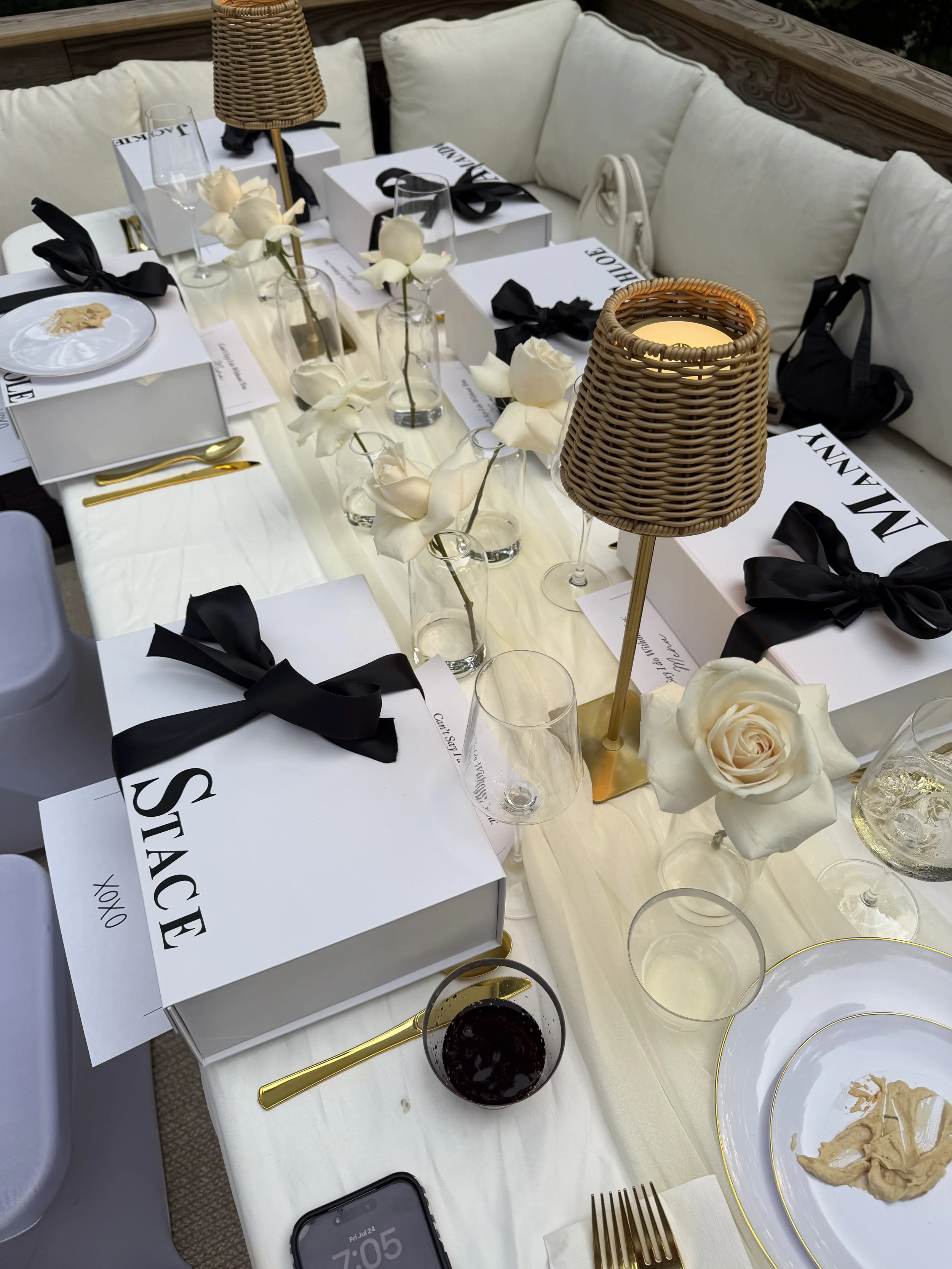 A dining table decorated with white roses in glass vases, black bows on white boxes labeled "STACHE," gold cutlery, and a welcome lamp. There are also empty wine glasses, a plate with some food, and a smartphone on the table. The seating includes cus