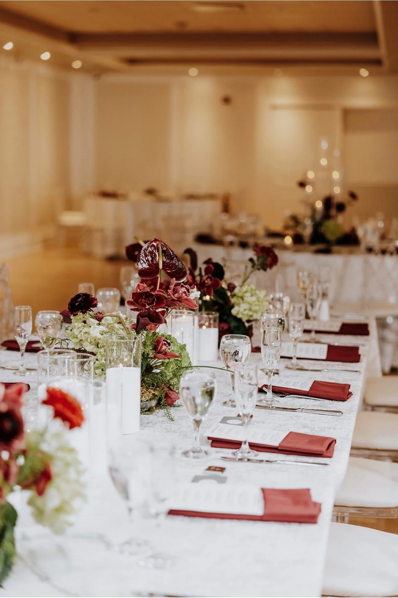 Elegant banquet table decorated with burgundy and white floral arrangements, surrounded by glassware, candles, and burgundy napkins, set for a formal event in a warmly lit room.