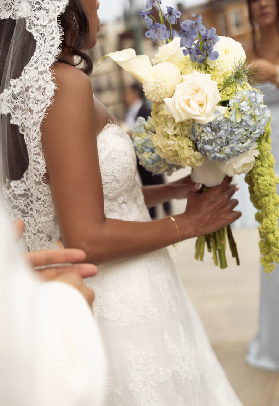 A bride holding a large bouquet of white and light blue flowers, wearing a lace wedding dress and veil, during a wedding ceremony.