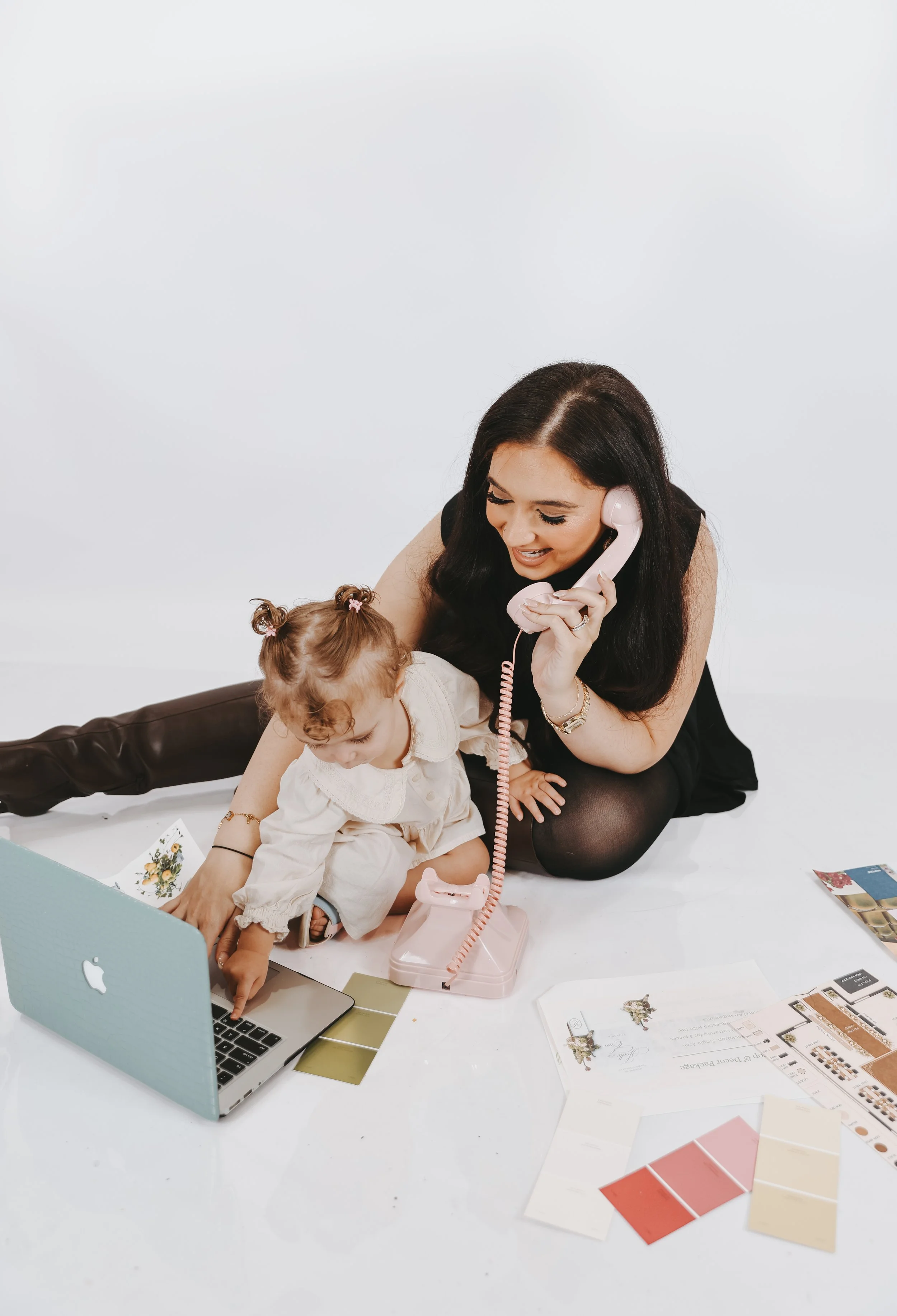 A woman and a young girl sitting on a white floor, with the woman talking on a pink vintage telephone and the girl pointing at a small laptop computer. The table is scattered with papers, color swatches, and brochures.