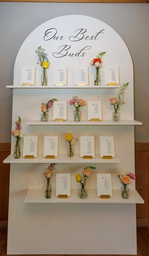 A decorative display titled 'Our Best Buds' featuring small flower arrangements in glass vases, each paired with a poem or note on a card, arranged on white shelves in a tiered display.