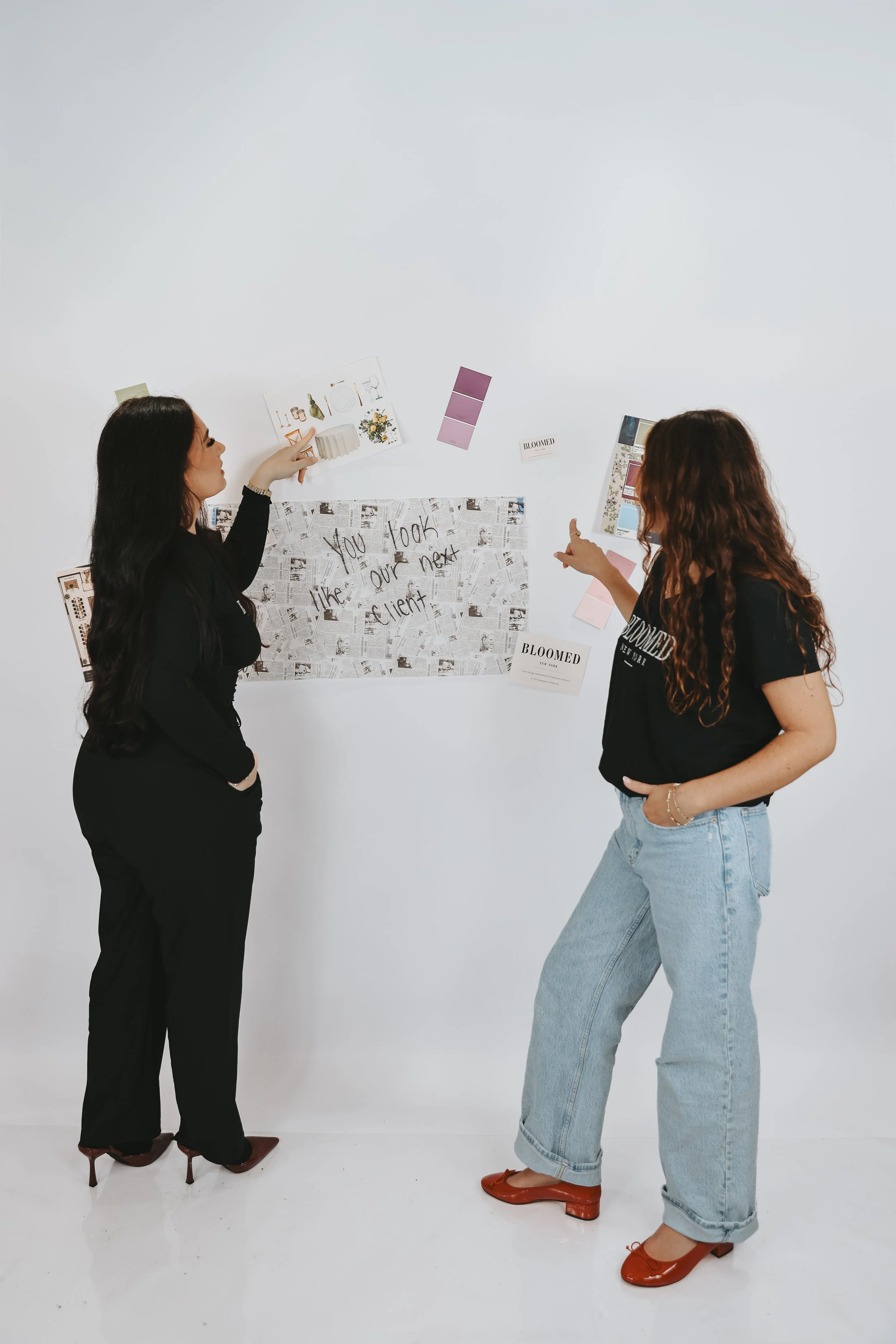 Two women standing in front of a white wall, engaging in a discussion about a bulletin board with various notes and images. One woman has long black hair, is dressed in black, and is pointing at the board. The other woman has long curly brown hair, i