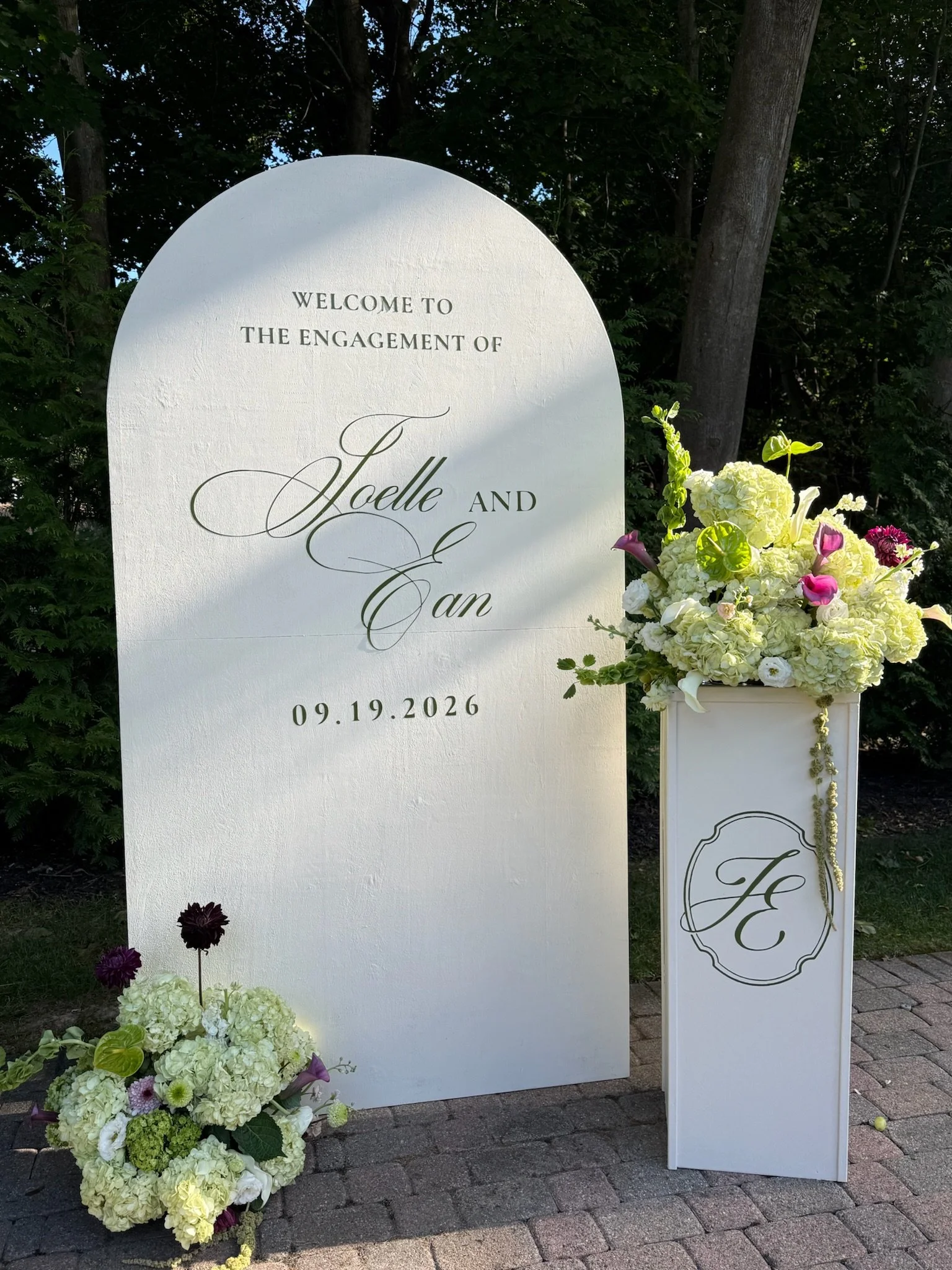 A white wedding sign with floral arrangements, welcoming guests to an engagement celebration for Joelle and Ian on September 19, 2026, set outdoors with trees in the background.