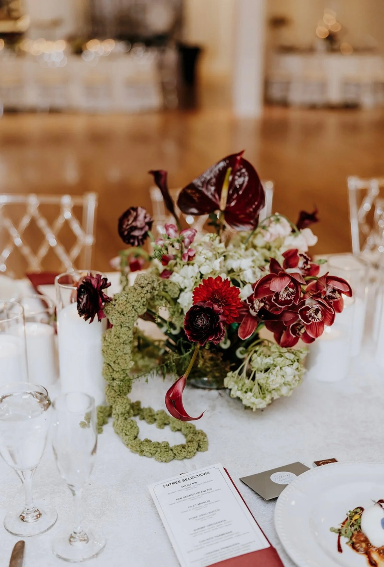 A floral centerpiece on a table at an indoor event with a menu, glasses, and plates visible in the foreground.