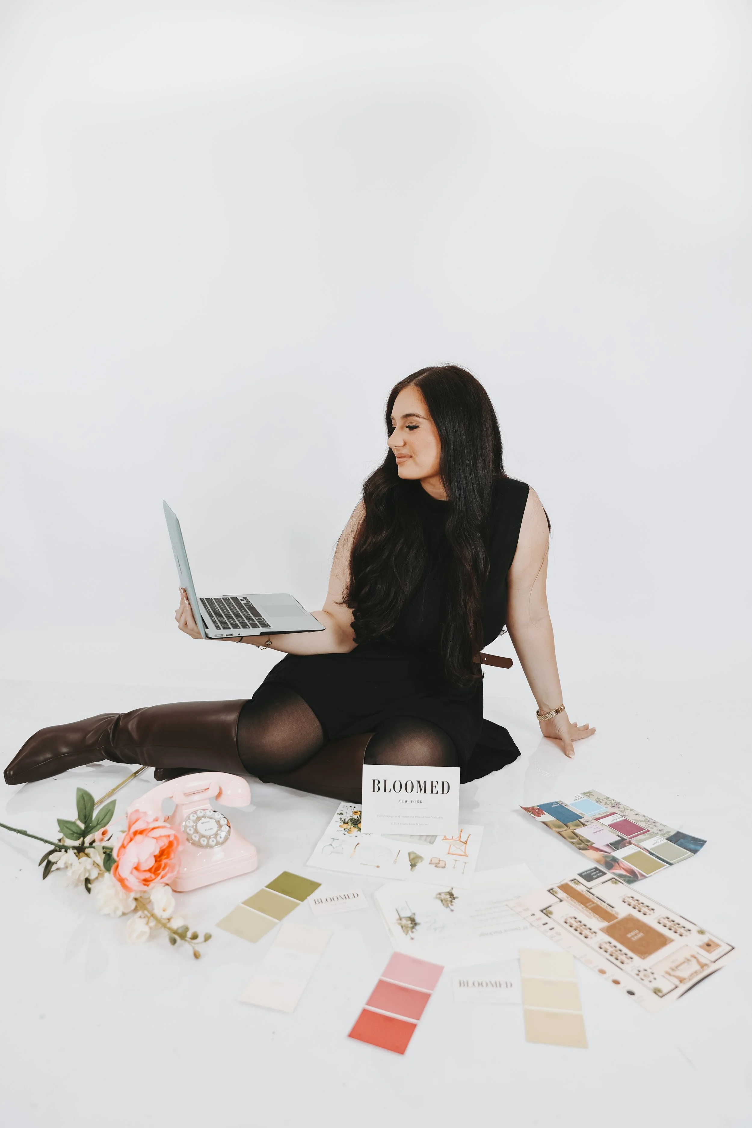 A woman with long dark hair wearing a black dress and brown knee-high boots is sitting on the floor with a laptop, surrounded by fashion magazines, color swatches, and a floral arrangement, with a white background.