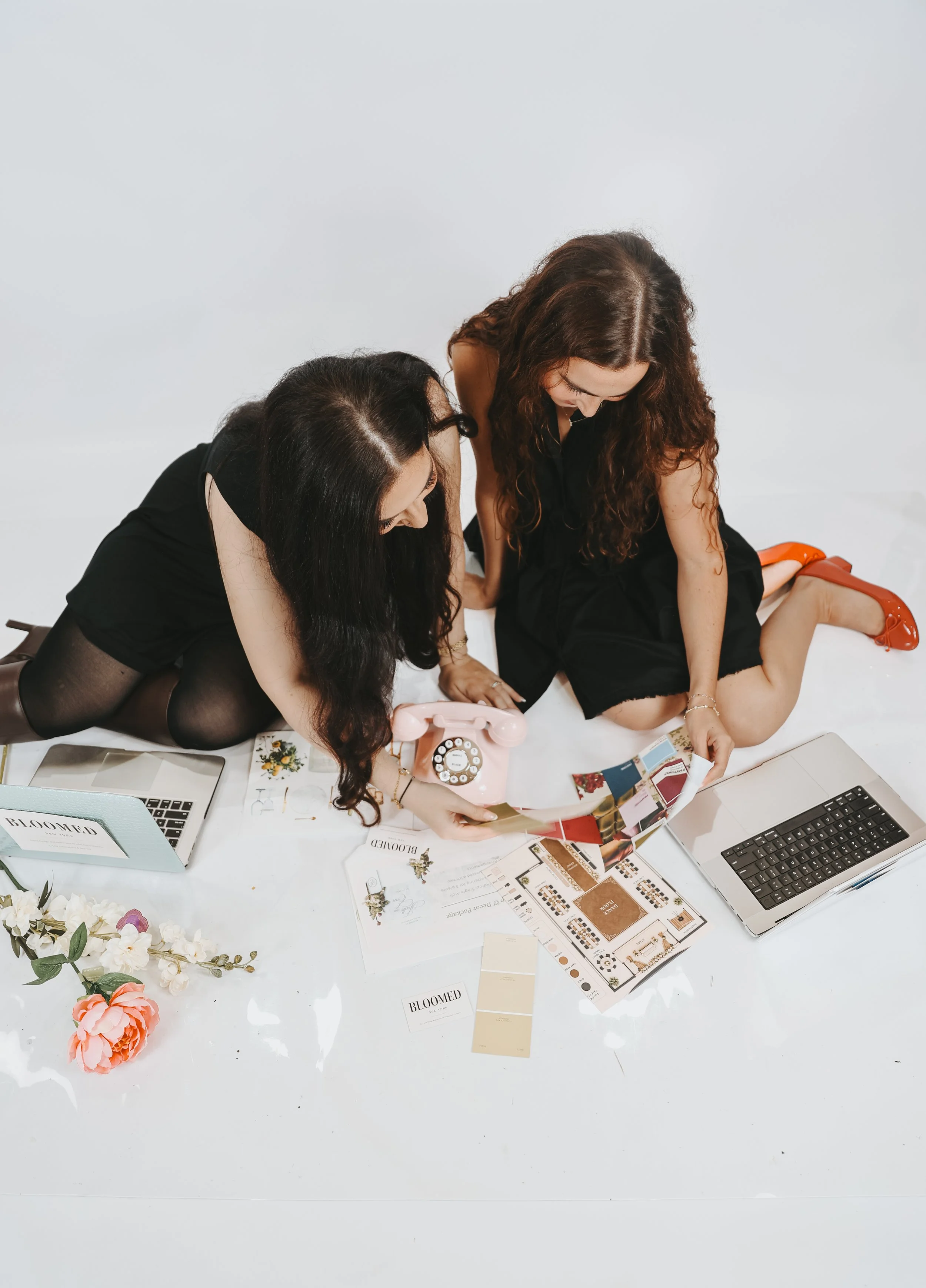 Two women laying on a white table surrounded by floral arrangements, color swatches, magazines, a pink rotary phone, and laptops, engaging with fashion and interior design materials.
