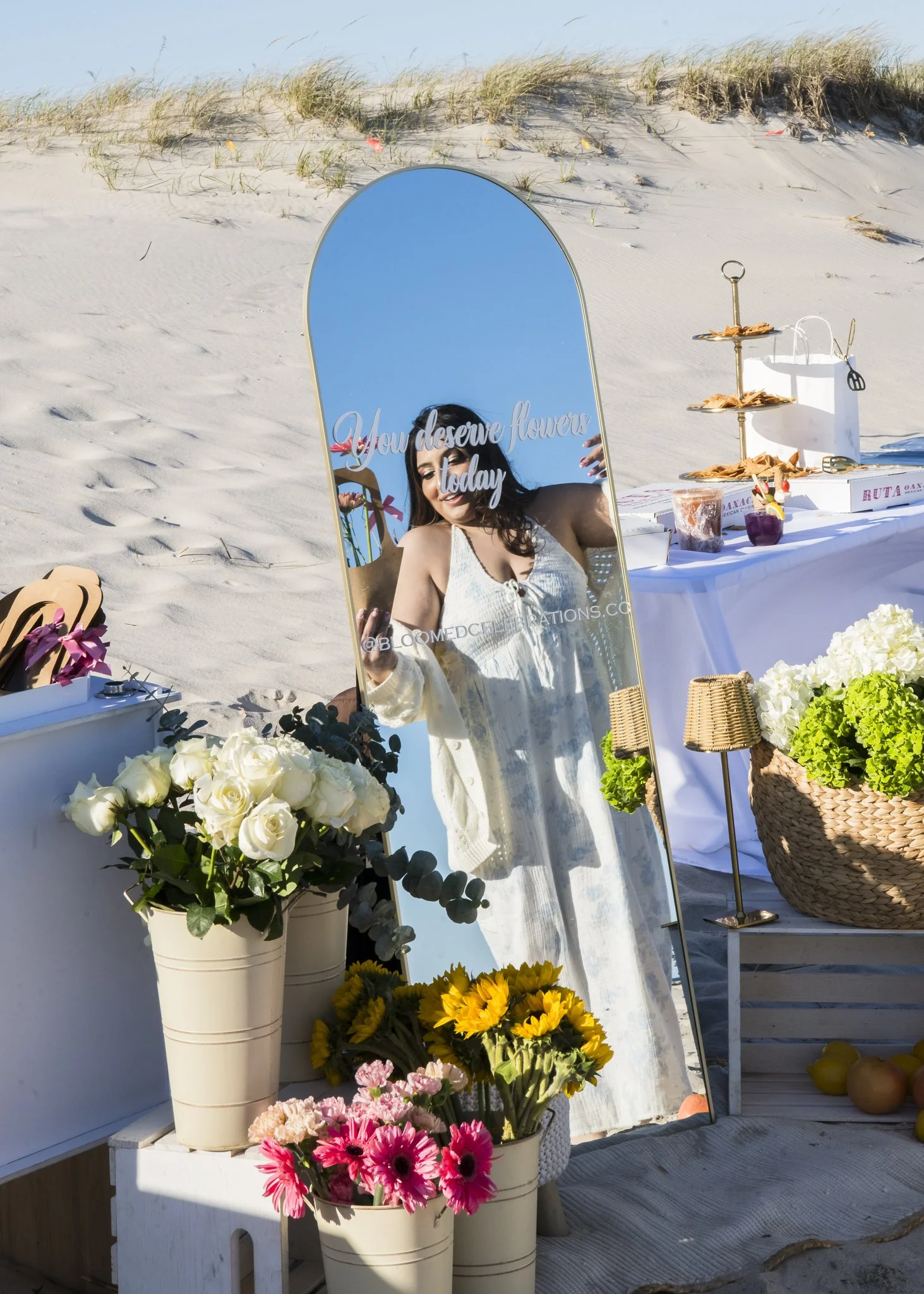 A beachside flower stand with various colorful flowers, a mirror with the words "You deserve flowers today," and a table with decorative items and fruit in the sand.