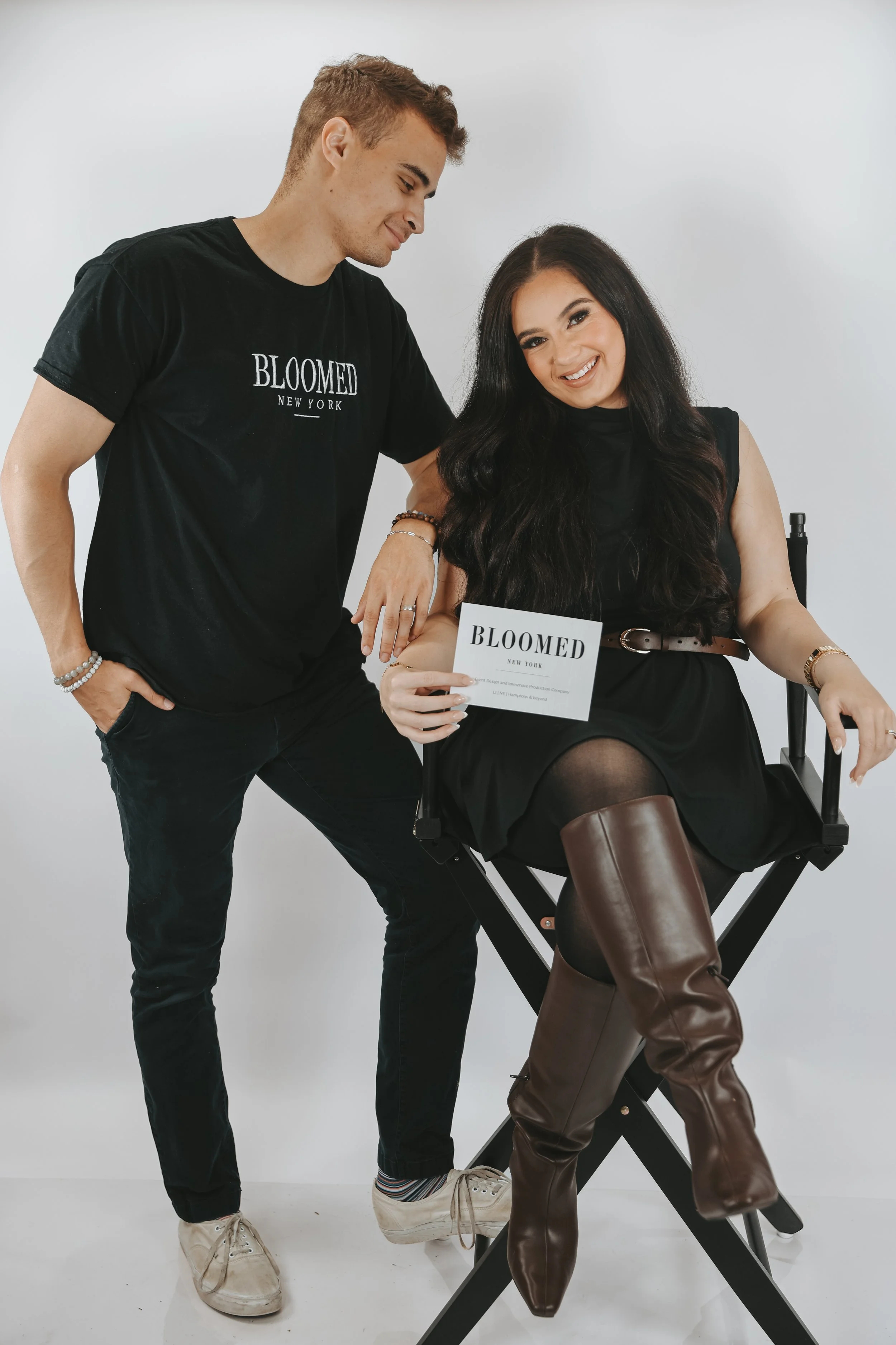 A man and woman posing together in front of a plain white background. The woman is seated on a director's chair with a sign that reads 'BLOOMED NEW YORK,' wearing a black dress and brown thigh-high boots, and smiling at the camera. The man is standing next to her, leaning slightly, wearing a black t-shirt with 'BLOOMED NEW YORK' printed on it, and looking at her with a smile.