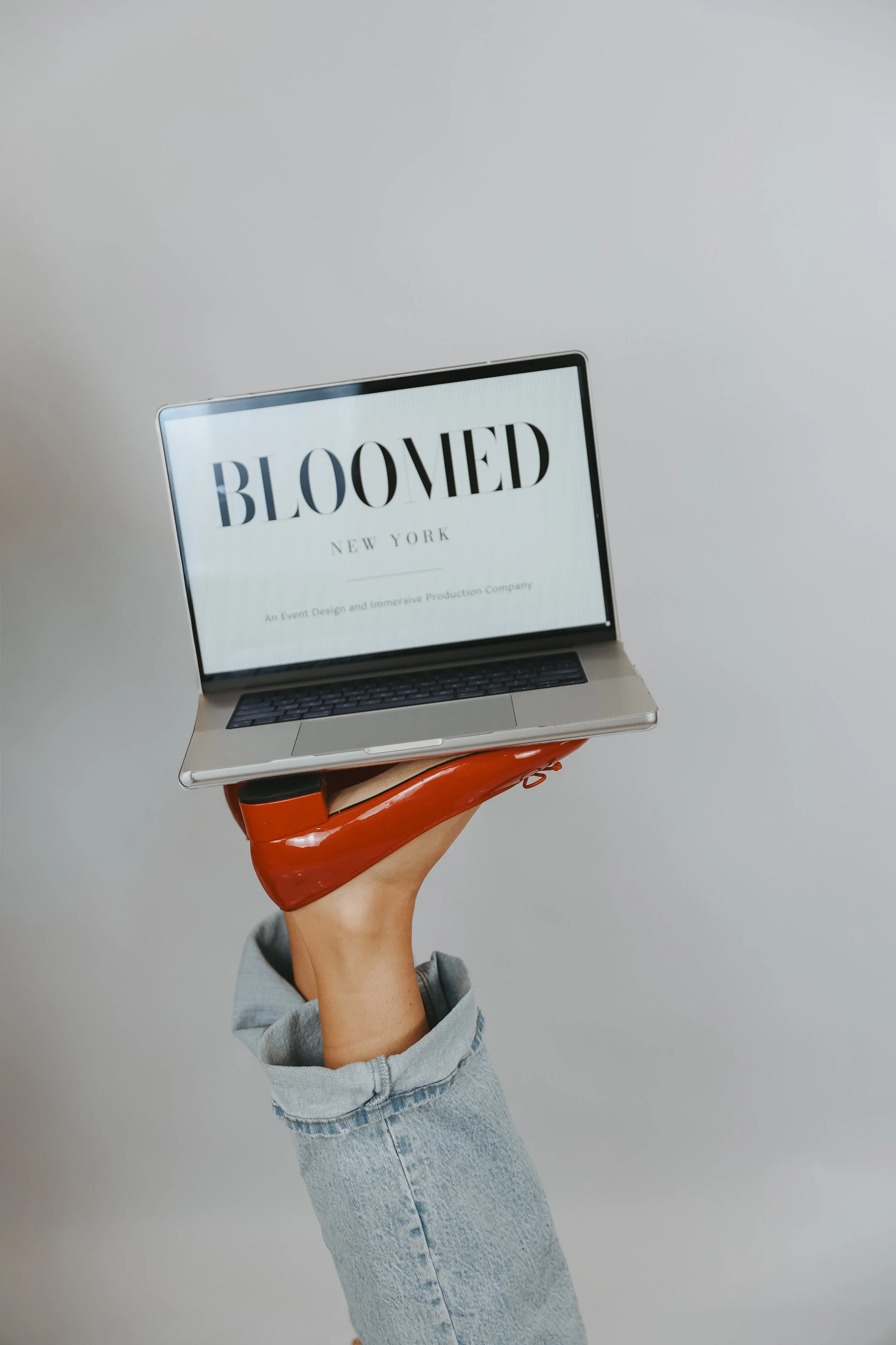 A person holding a laptop with the screen displaying the text 'BLOOMED NEW YORK' and the subtext 'An Event Design and Immersive Production Company' against a plain, light gray background.