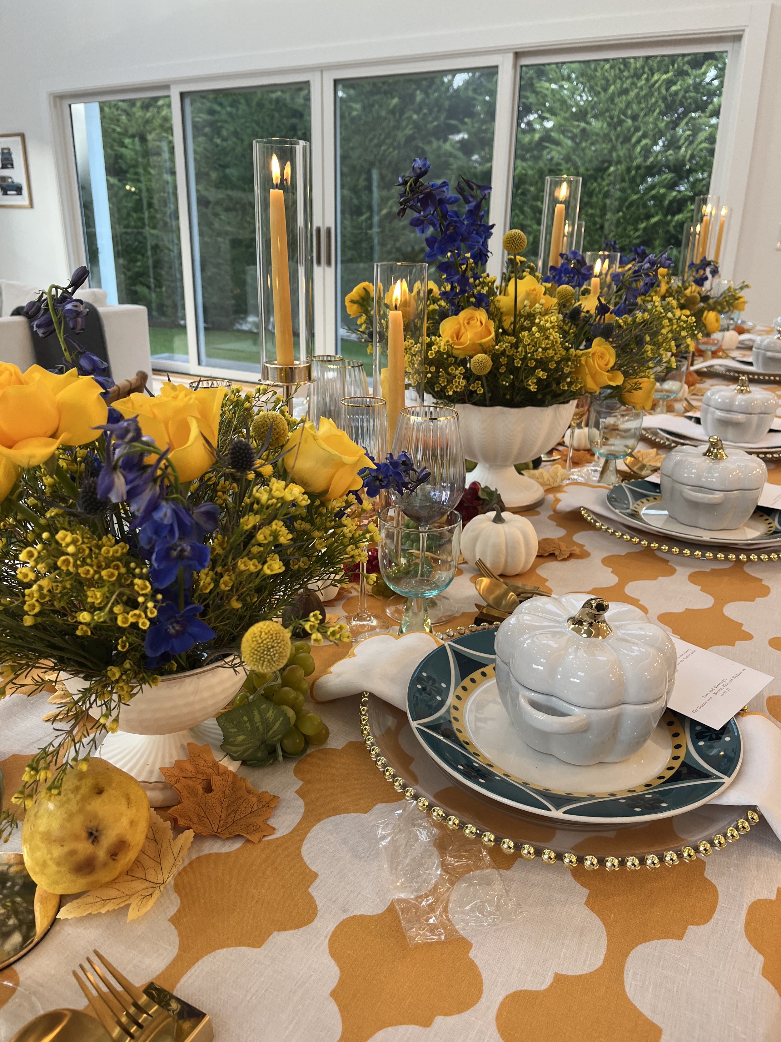 A decorated dinner table with floral centerpieces of yellow roses and purple flowers, tall candles in glass holders, pumpkin-shaped dishes, and festive fall leaves and fruit, set near large sliding glass doors revealing a green outdoor scene.