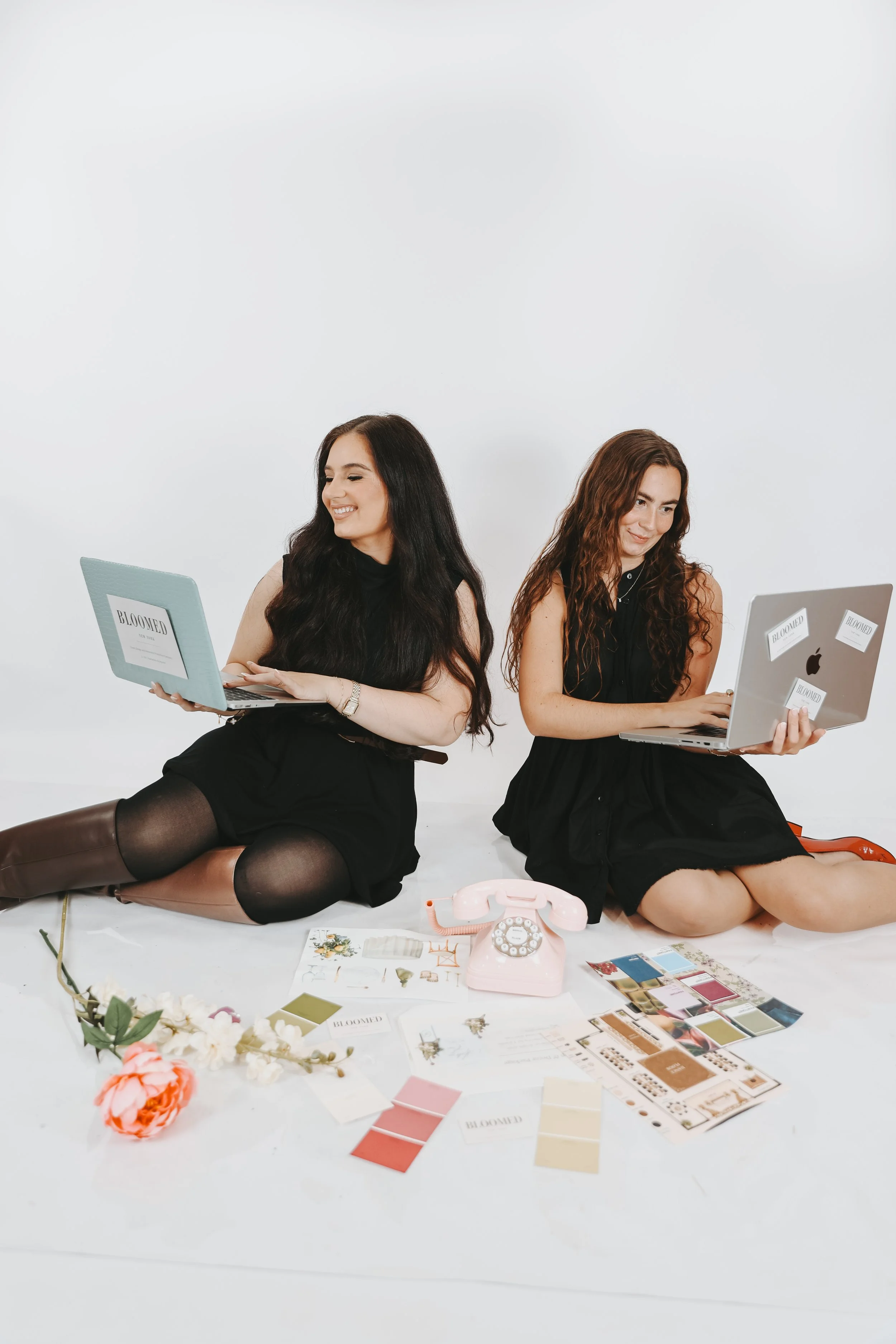 Two women sitting on the floor with laptops, surrounded by flowers, color swatches, and decorating samples, in a minimalist photography studio with a plain white background.