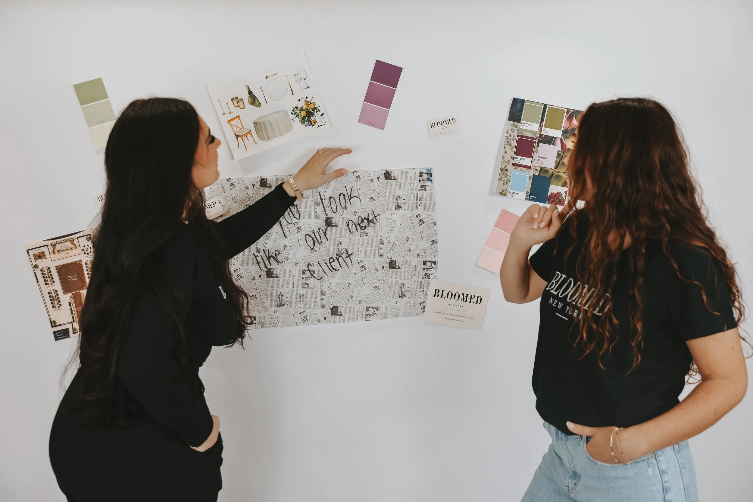 Two women are discussing design ideas on a wall covered with swatches and images, with one woman pointing to a picture of a table and flowers, and the other listening with her hand on her chin.