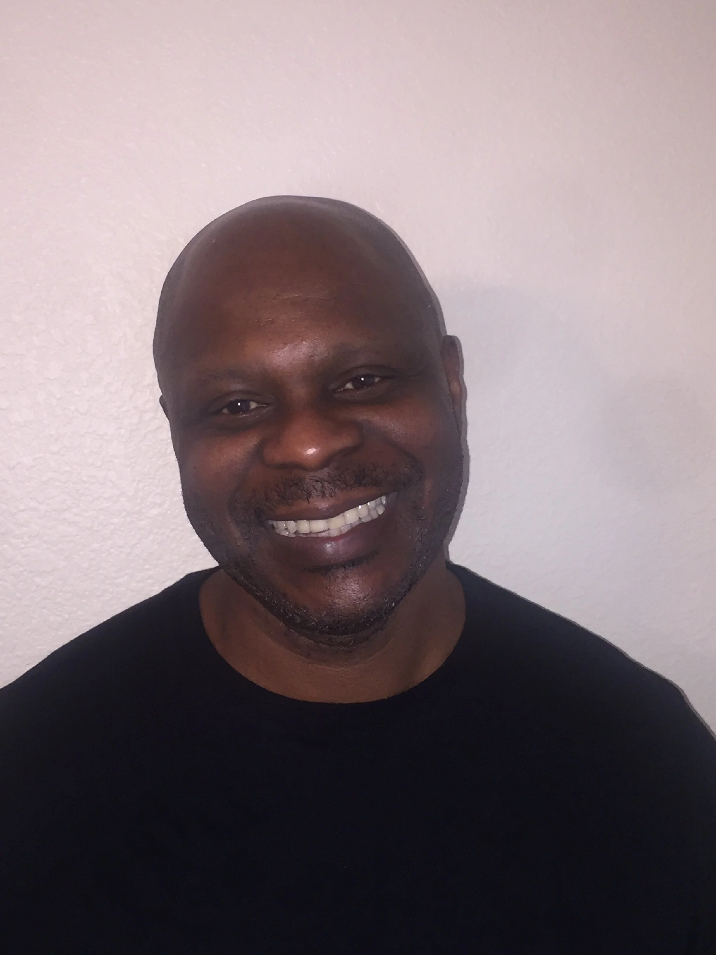 Smiling man with a close-cropped haircut, wearing a black shirt, standing against a plain white wall.