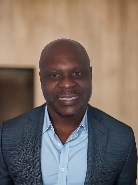 A smiling African American man with a bald head, wearing a light blue dress shirt and a dark blazer, standing indoors with a blurred background.
