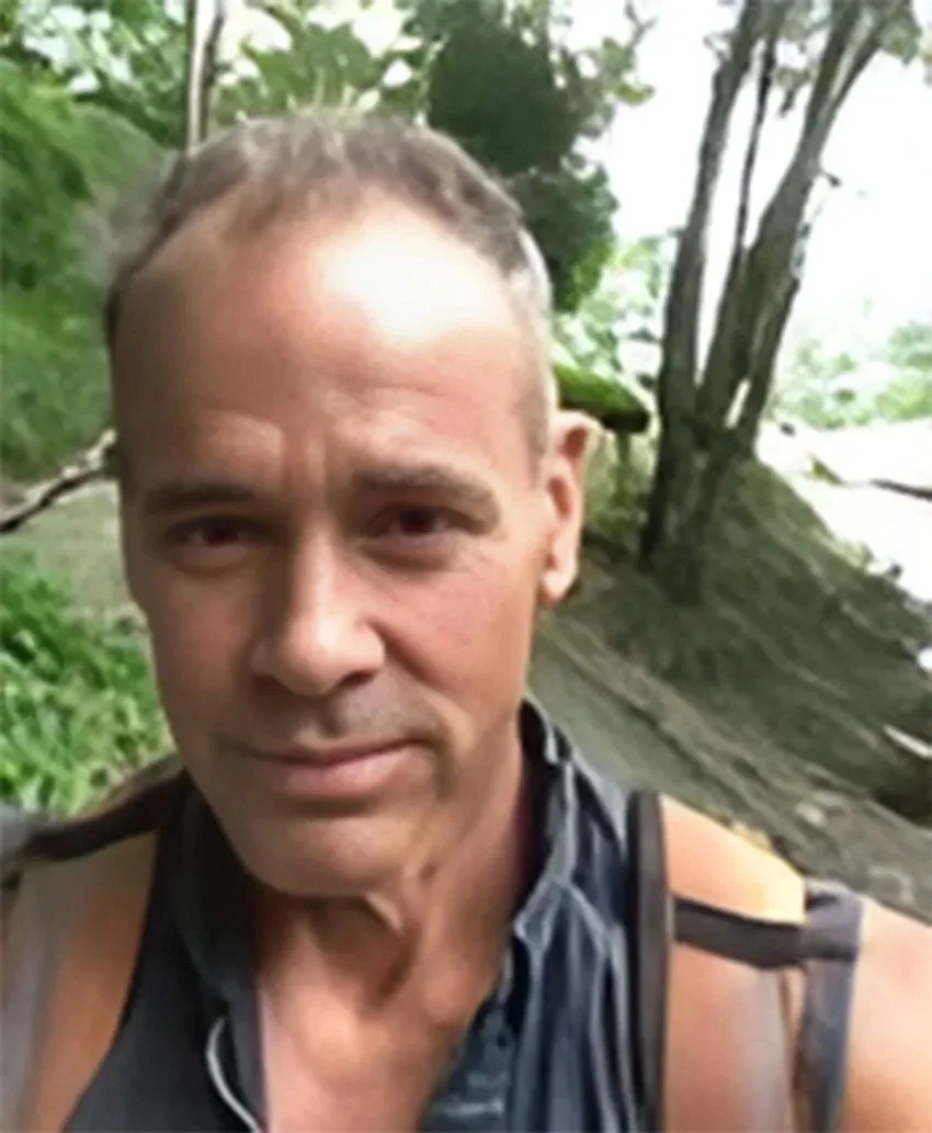 A man taking a selfie outdoors in a wooded area, wearing a black sleeveless shirt, with trees and a shoreline in the background.