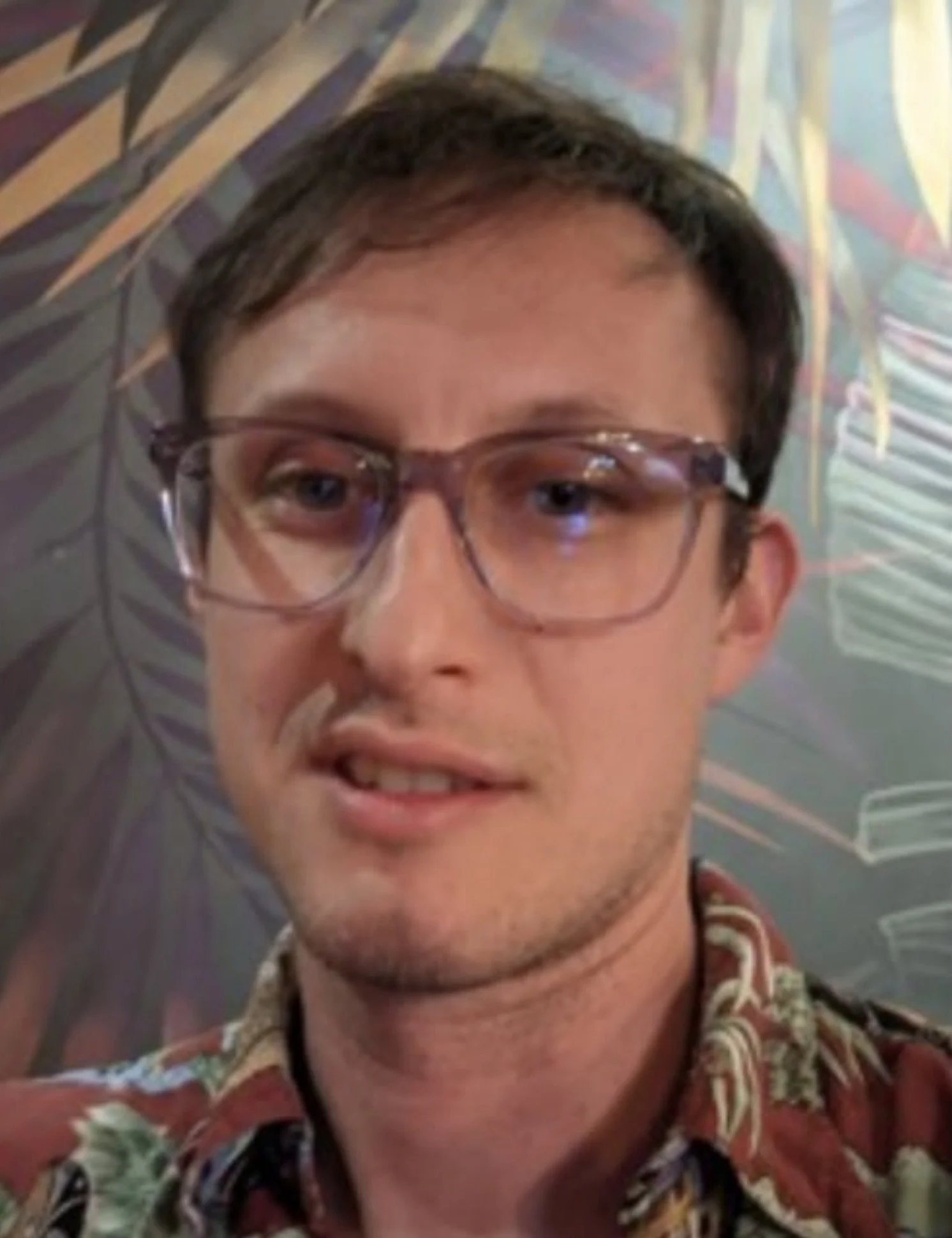 Close-up of a young man with brown hair, wearing glasses and a colorful, patterned shirt, standing in front of a multicolored background.