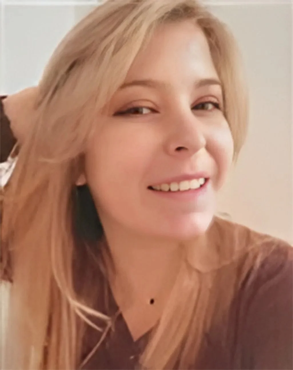 Close-up of a smiling woman with blonde hair, wearing a dark top, in a well-lit indoor setting.