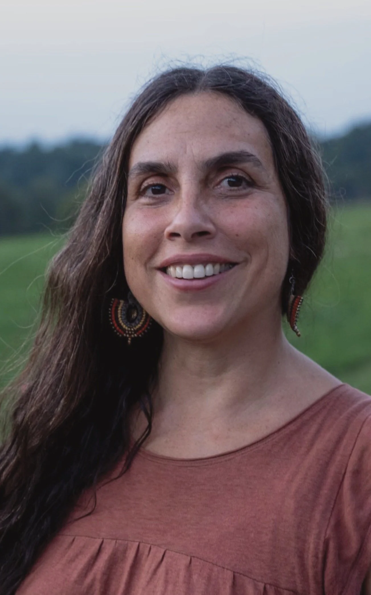 A young woman with long dark hair, wearing large beaded earrings and a reddish-brown top, smiling outdoors with a green field and cloudy sky in the background.