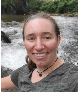 Woman with braided hair smiling near a river with trees in background.