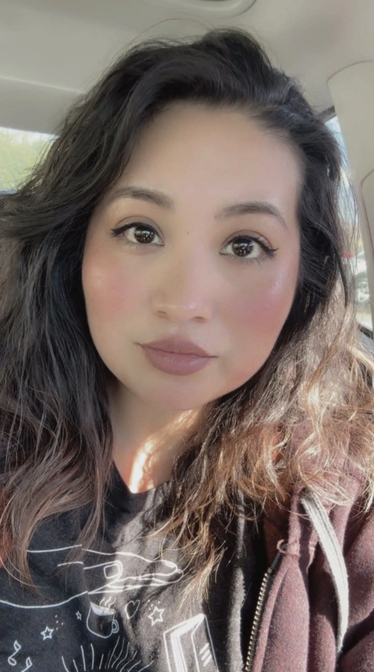 A young woman with long, wavy, dark hair, wearing makeup with eyeliner and lipstick, sitting inside a vehicle.