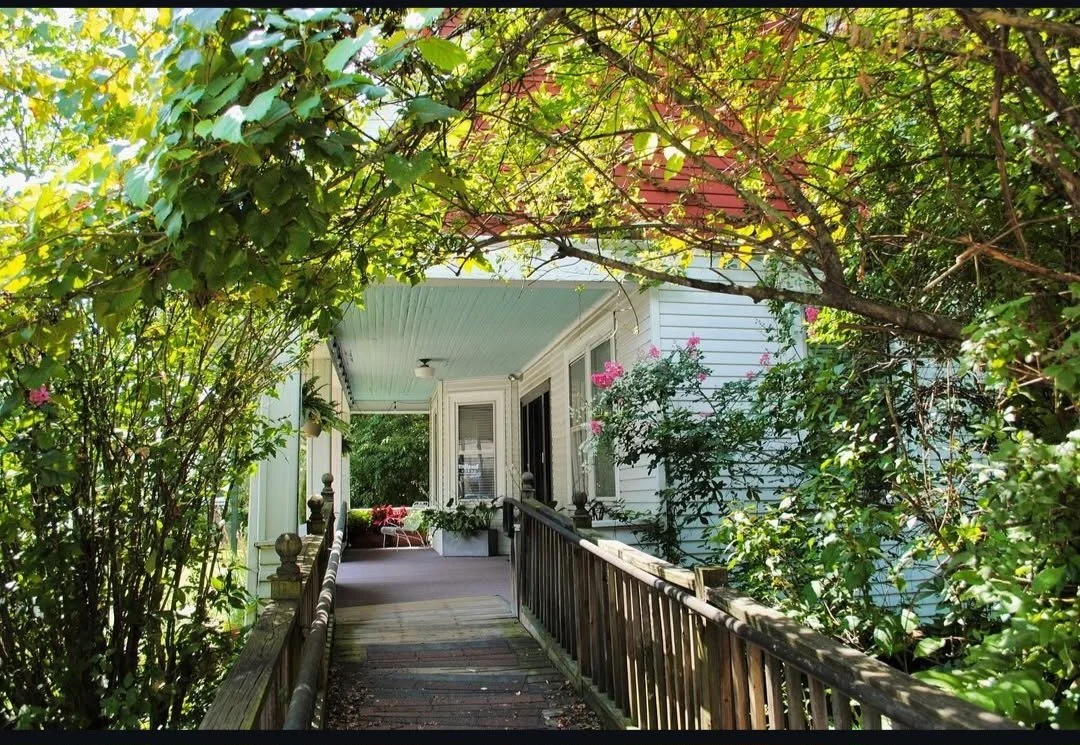 A porch surrounded by greenery and flowering bushes, with a wooden railing and a house in the background.
