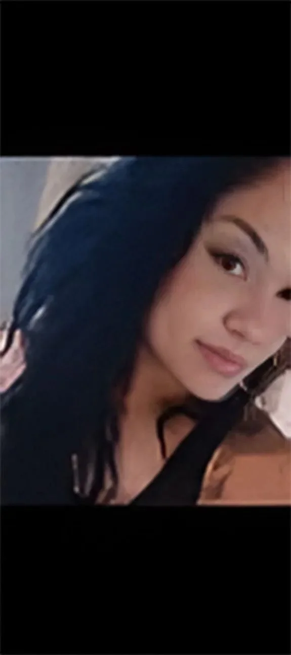 Close-up of a woman with long dark hair and light skin, smiling softly. She appears to be wearing a black top and is in an indoor setting.