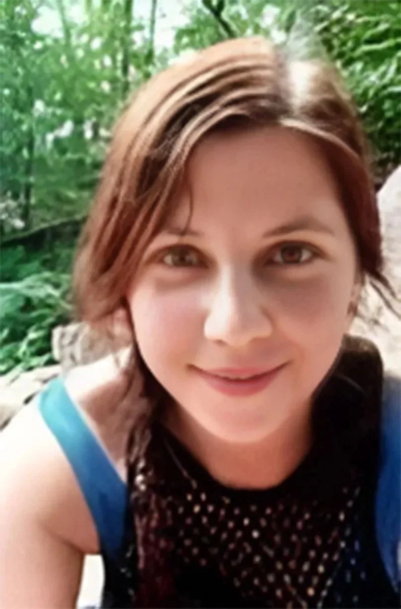 Close-up of a smiling woman with brown hair and brown eyes, outdoors in a green, wooded area.