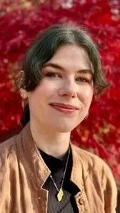 Young woman outdoors with red autumn leaves in the background, smiling, wearing a brown jacket and a necklace.