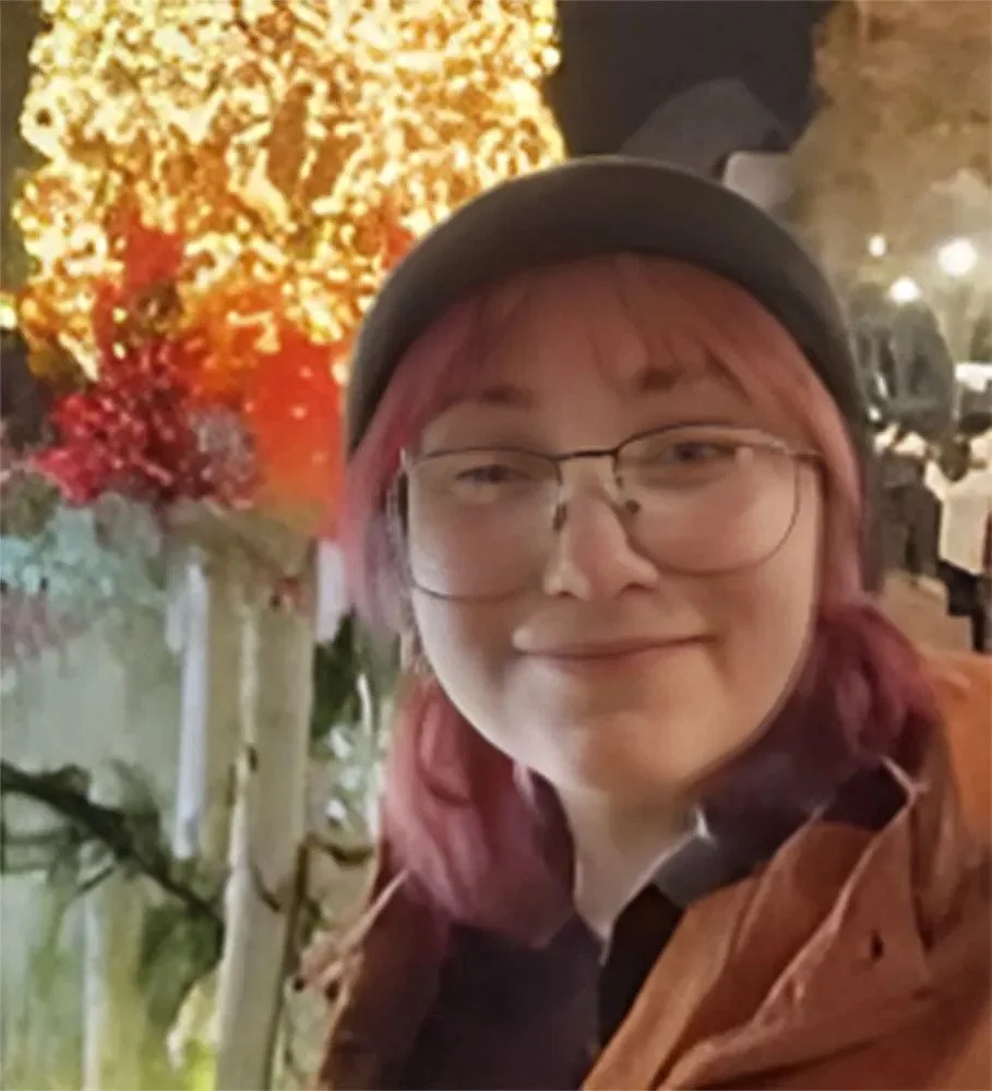Person with pink hair, glasses, and a black hat smiling outdoors at night, with yellow and red lit trees or decorations in the background.