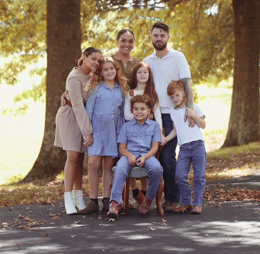 A family of seven posing outdoors in autumn, standing on a paved path with large trees and fallen leaves in the background.