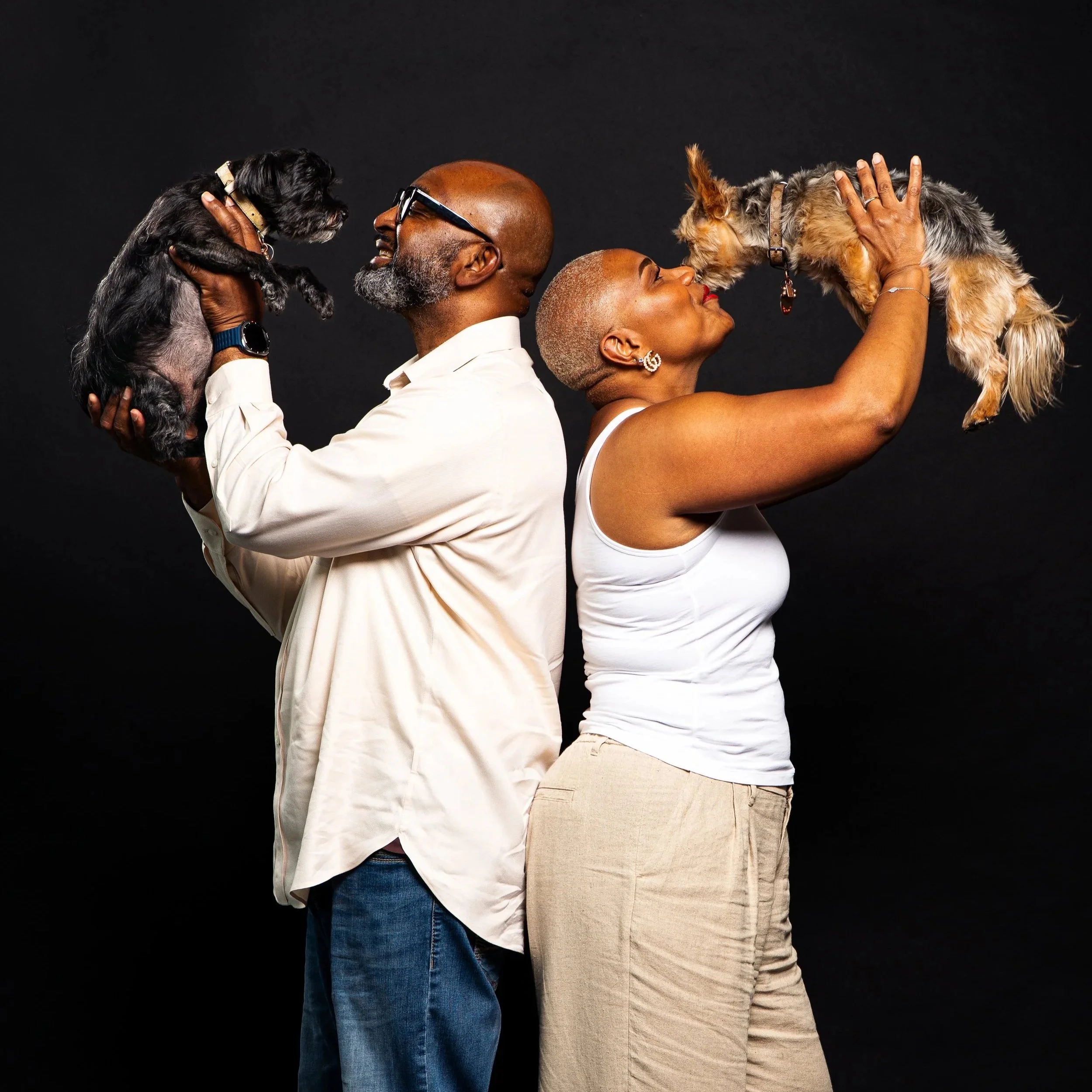 A man and woman are holding dogs up to each other's faces, both smiling against a black background.