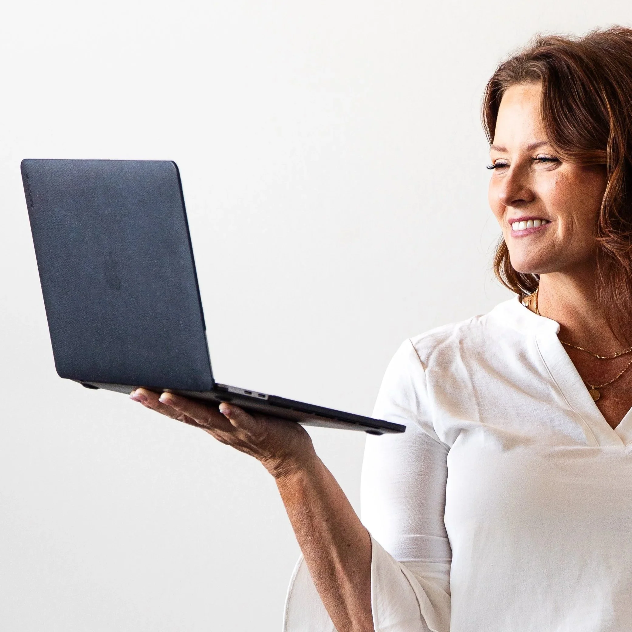 A woman with brown hair, wearing a white shirt, smiling while looking at a black laptop.