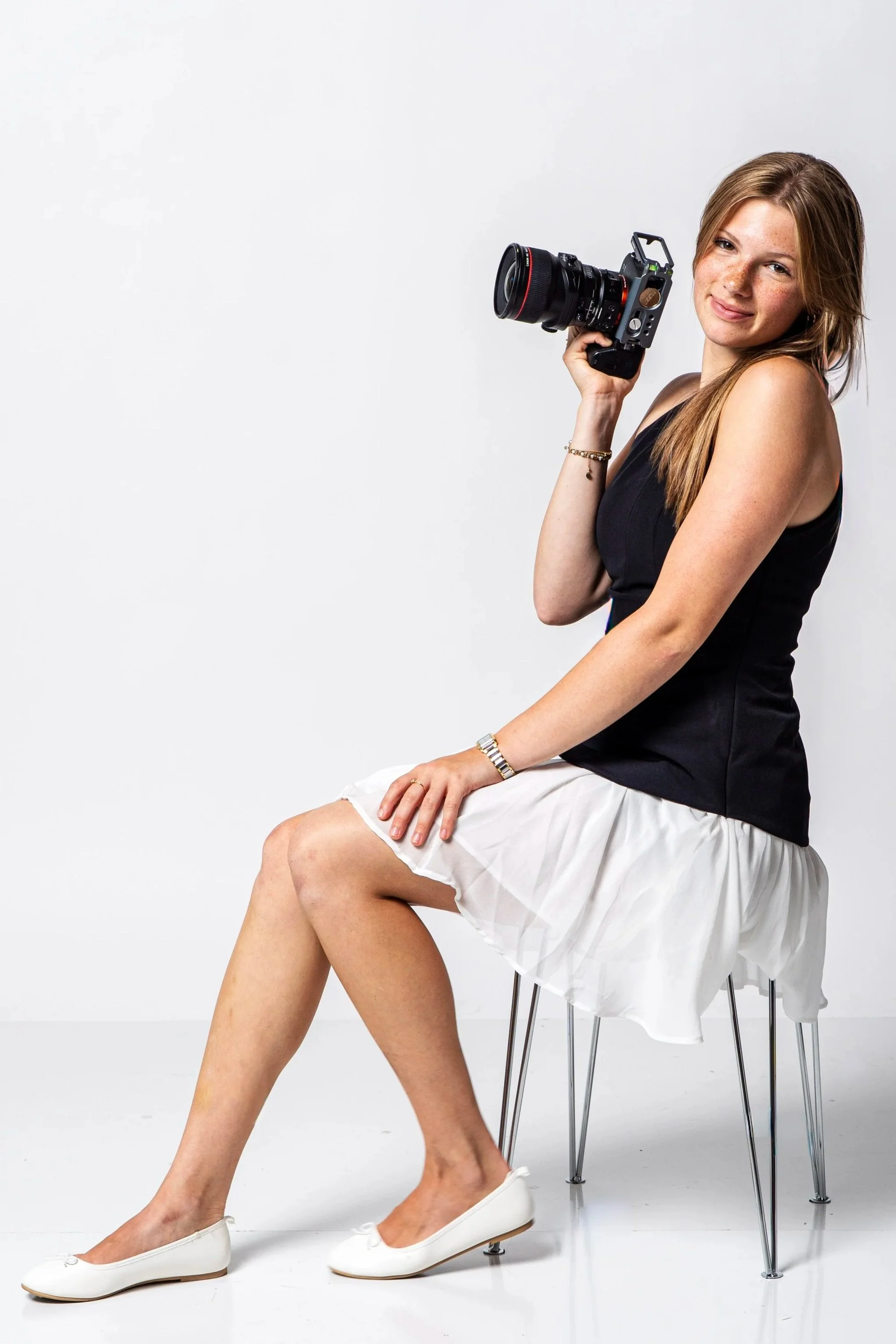 A young woman with long, light brown hair and freckles sitting on a clear chair, holding a professional camera in her right hand, smiling at the camera, wearing a black top, a white skirt, and white flats, against a plain white background.