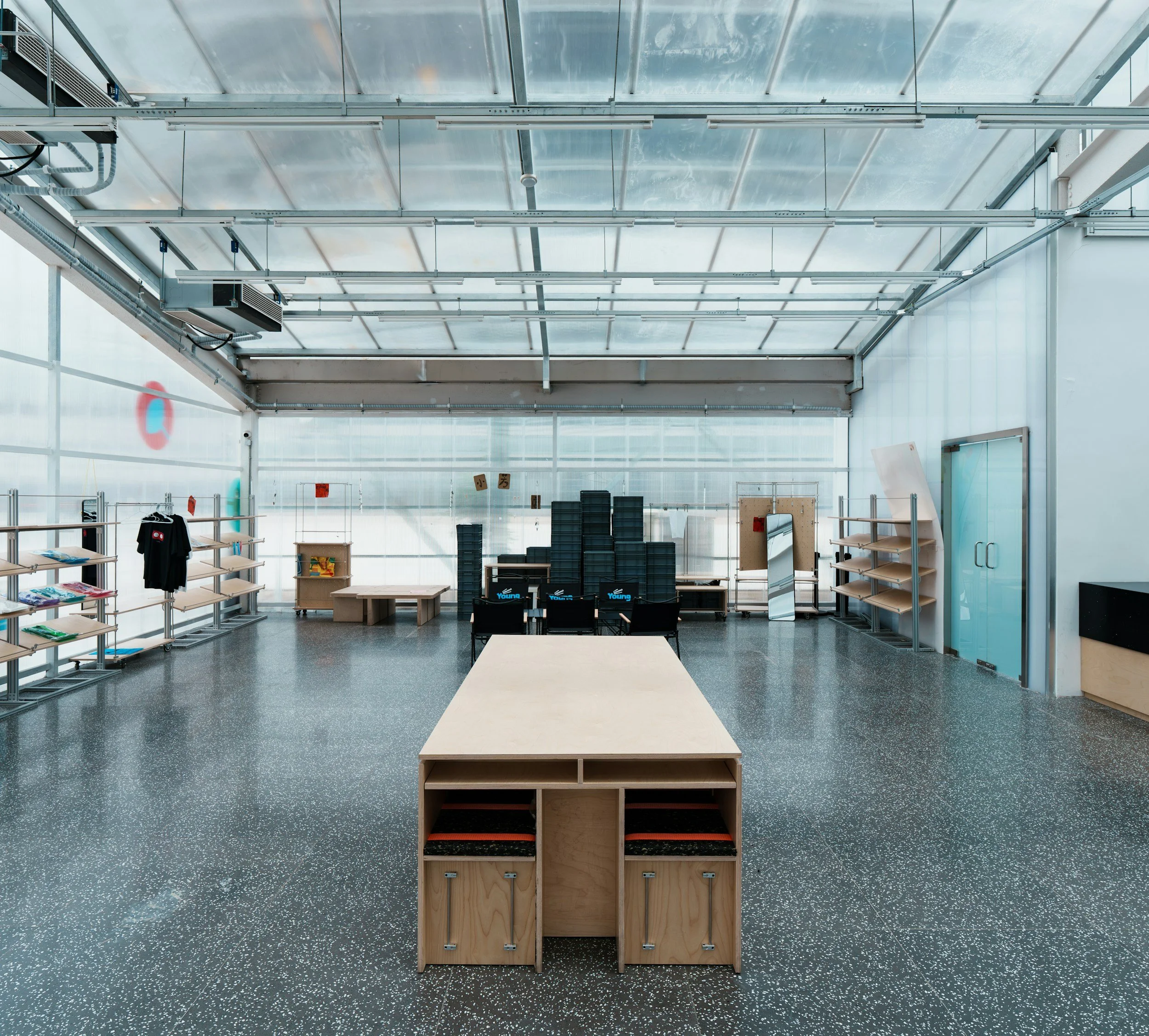 Interior of a modern store or exhibition space with empty wooden display shelves on both sides, a central wooden display unit, and black chairs with blue logos. The ceiling has a glass roof with visible metal supports and electrical conduit.