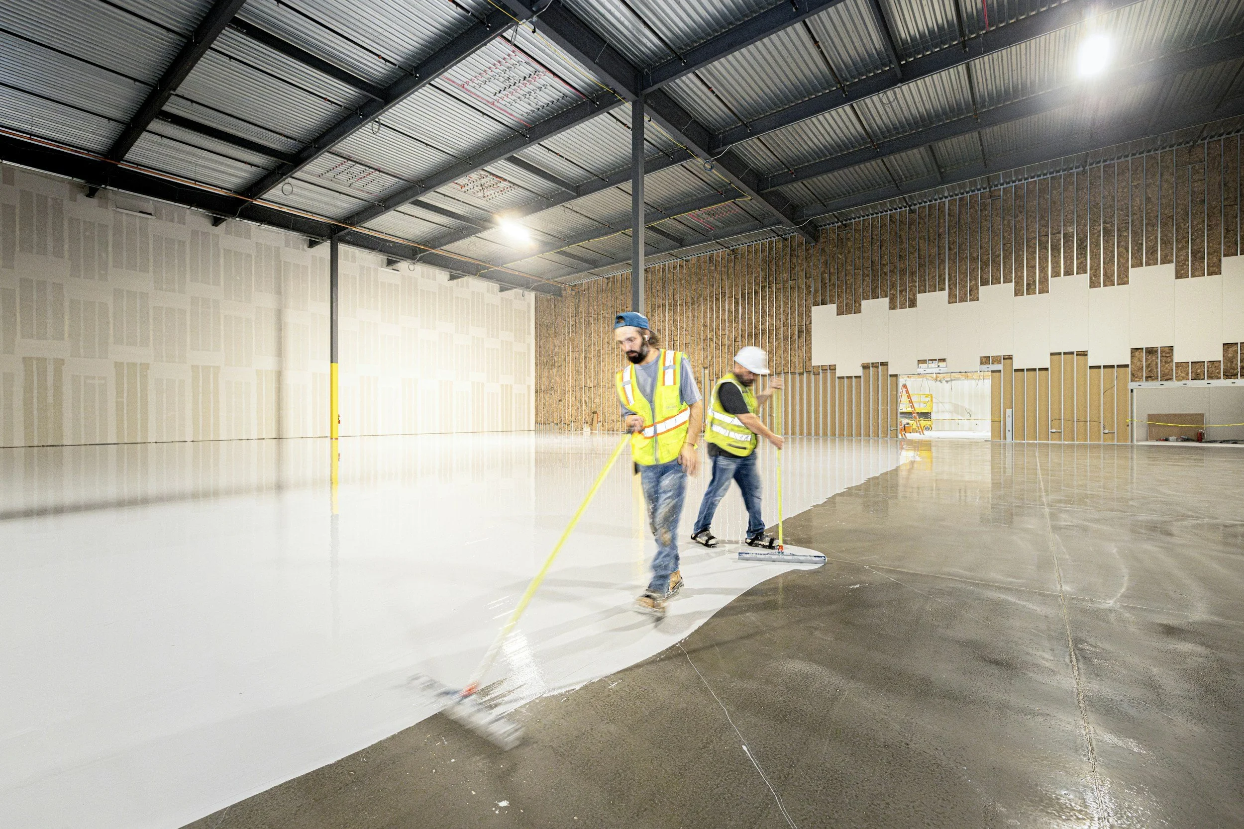Two construction workers wearing safety vests and helmets are applying epoxy coating to a concrete floor inside a large building under construction.
