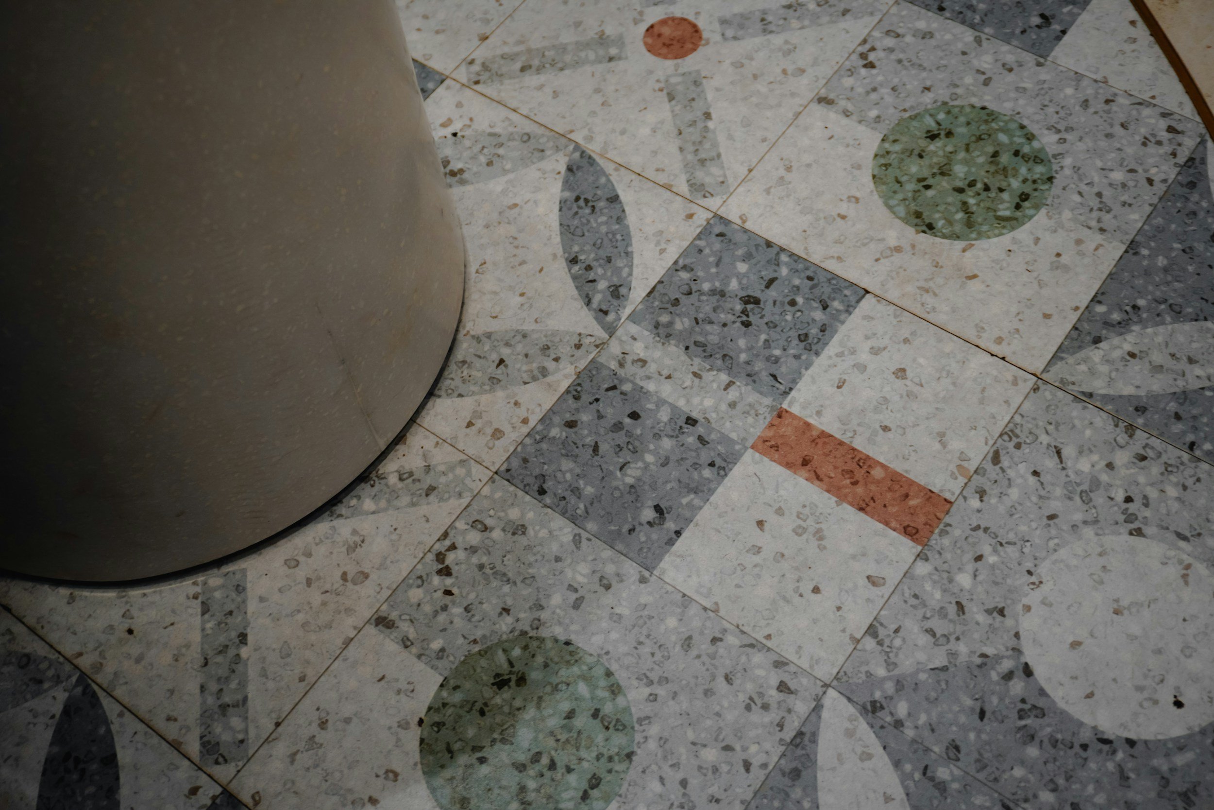 Close-up of terrazzo flooring with various colored circular and geometric patterns, next to a curved metallic object on the left.