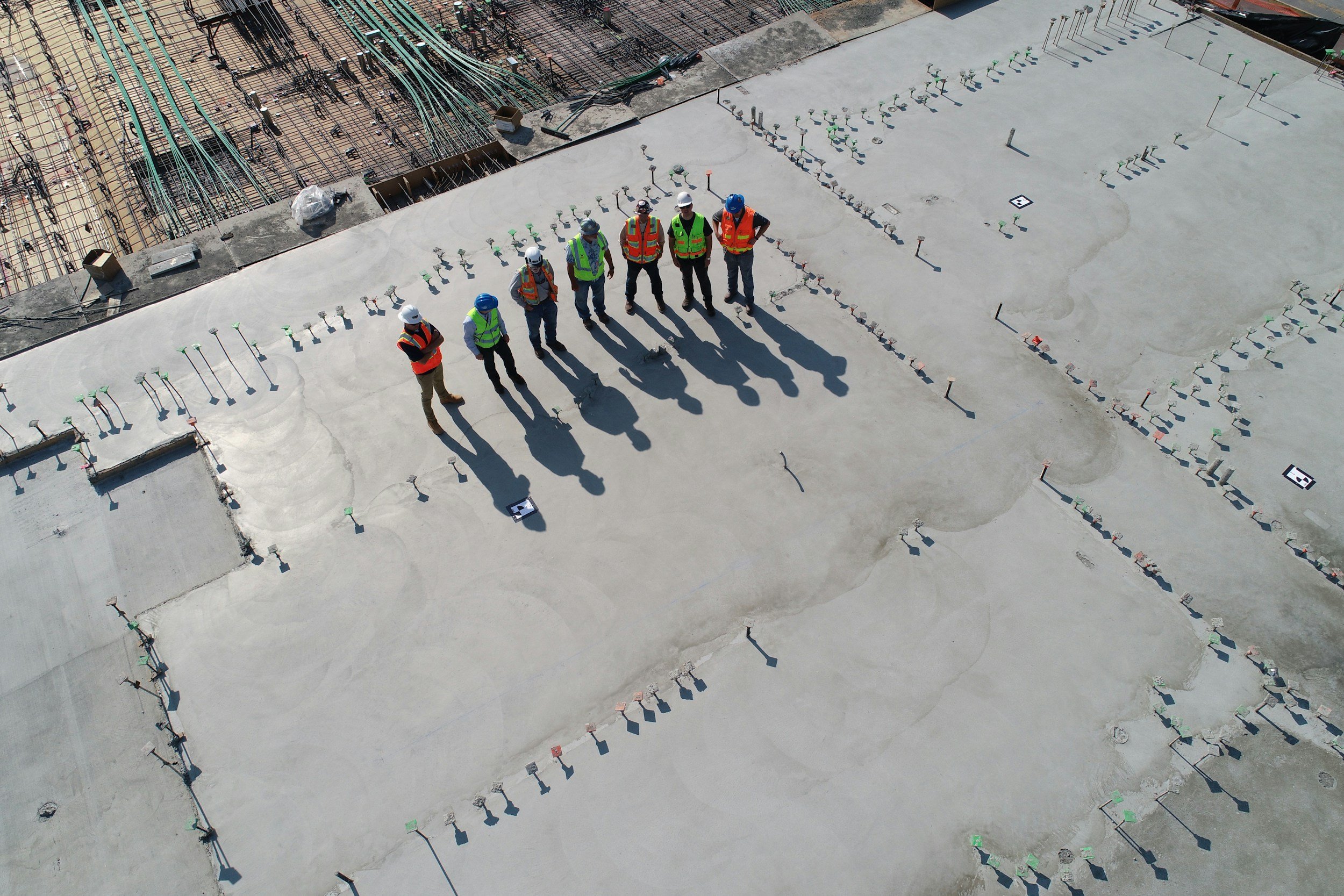 Six construction workers wearing hard hats and safety vests standing on a concrete slab at an outdoor construction site, with partially completed building structure and rebar in the background.